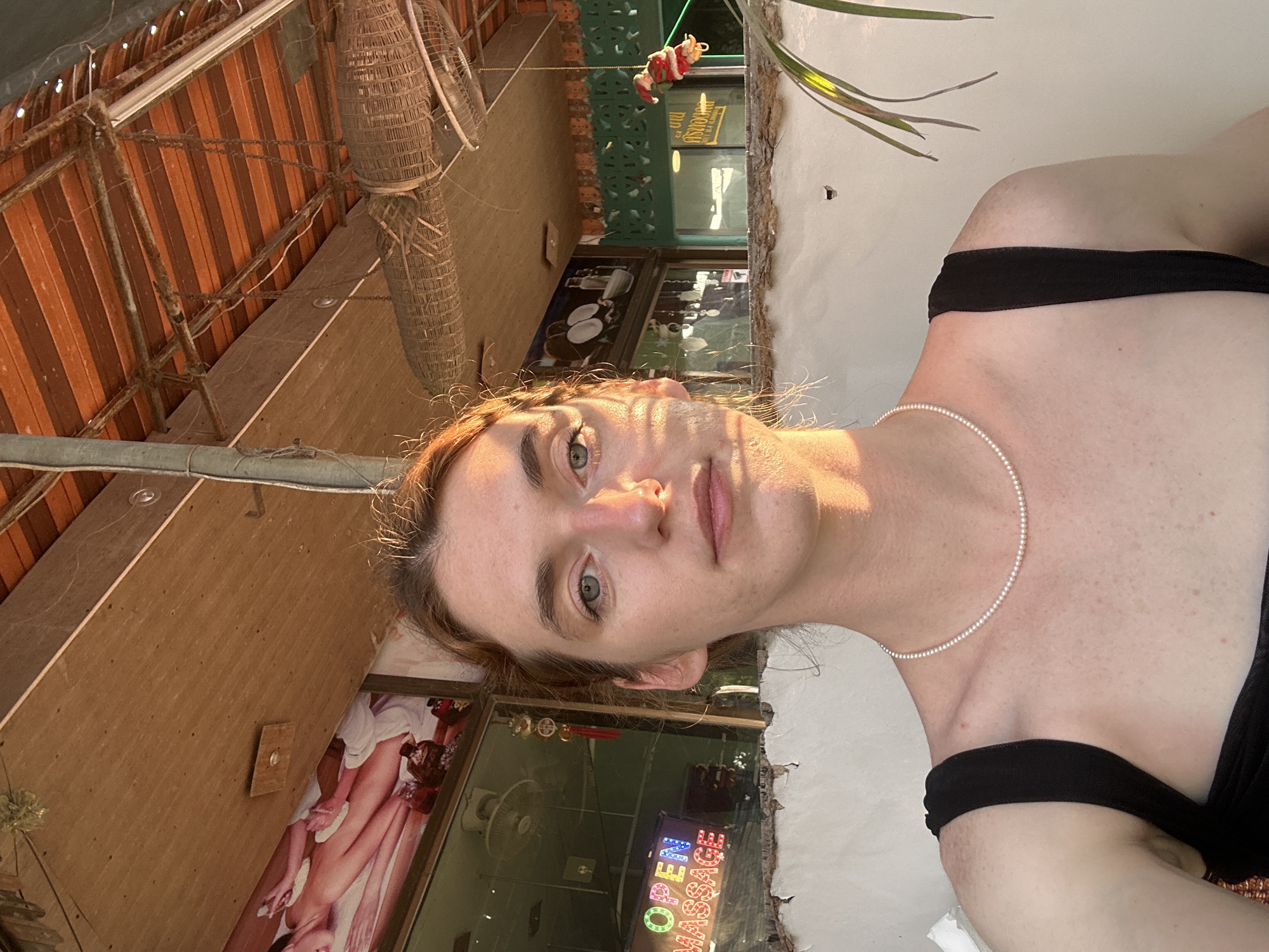 A selfie taken in Baan Bang Rak, Thailand shows a person with fair skin, light brown hair, and a pearl necklace wearing a black top. The person is in a pool. Behind them, there is a building with a sign saying "Massage Open" in neon lights. The roof is visible in the background.