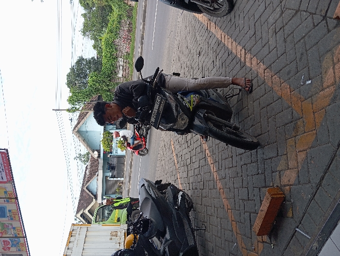 The scene depicts an outdoor urban environment in Legok, Indonesia, under an overcast sky. In the foreground, a young male with dark, curly hair, wearing a dark jacket, light-colored pants, and dark flip-flops, is positioned on a black scooter with license plate BM 2001 TGA. He is stationary, with his right foot on the grey interlocking paver ground and his right hand near his chin. Another large, dark scooter is parked to his left. A single red brick lies on its side on the paved ground, which features yellow chevron markings. In the midground, an asphalt road separates the foreground from the background. On the road, a male in a white shirt and light-colored helmet rides a red scooter from right to left. To the far left, a male wearing a green high-visibility vest stands near the green cab of a white container truck. Across the road, two single-story houses with light blue walls and brown roofs are visible behind fences. Dense green vegetation and trees are present behind the houses and along the right side of the road. A utility pole with overhead wires is visible against the sky. Partial commercial signage with red, white, and yellow elements is visible in the upper left corner.