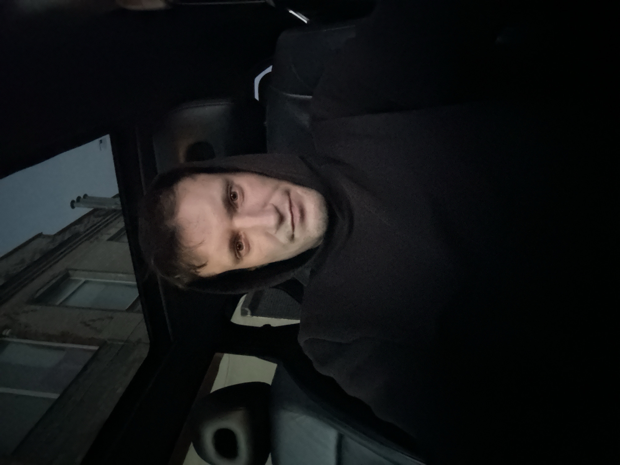 In Shatava, Ukraine, a man with short brown hair and a dark hoodie is captured in a selfie. He is seated inside a vehicle, with the interior of the car visible behind him. The car's black leather seats and a sunroof are discernible. Through the sunroof, a building with windows and a cloudy sky are visible, suggesting the photo was taken outdoors, possibly during twilight or a dimly lit environment. The man's facial expression is neutral.