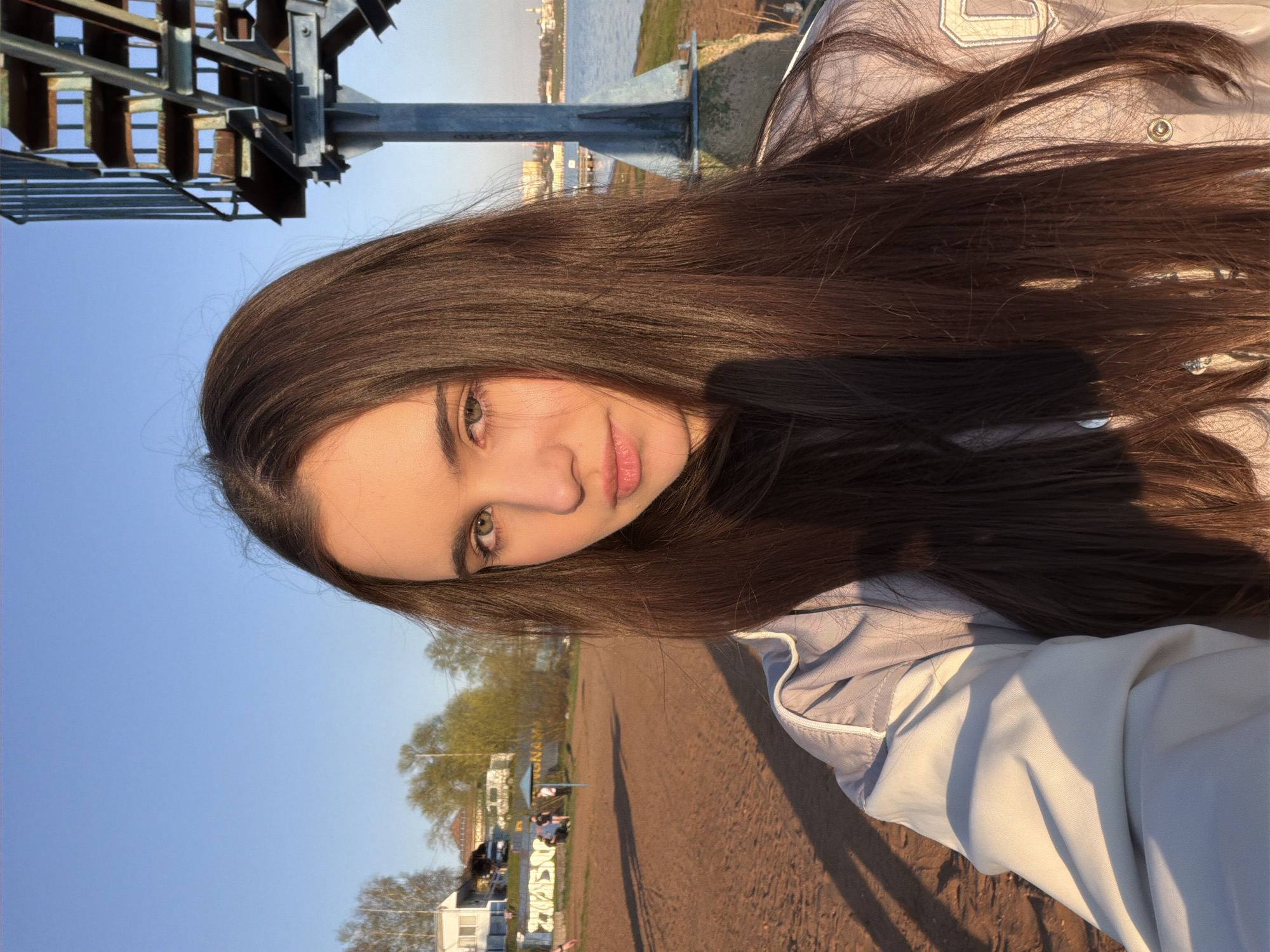 A young woman with long, dark hair is featured in a close-up shot, likely a selfie. She has fair skin, brown eyes, and light pink lips. The background includes a metal structure on the left, what appears to be a body of water in the distance, and a grassy area with buildings further back. The environment seems to be outdoors on a sunny day, with long shadows on the ground. Based on these observations, the photo was taken near the Izhevsk pond in the city of Izhevsk, Russia.