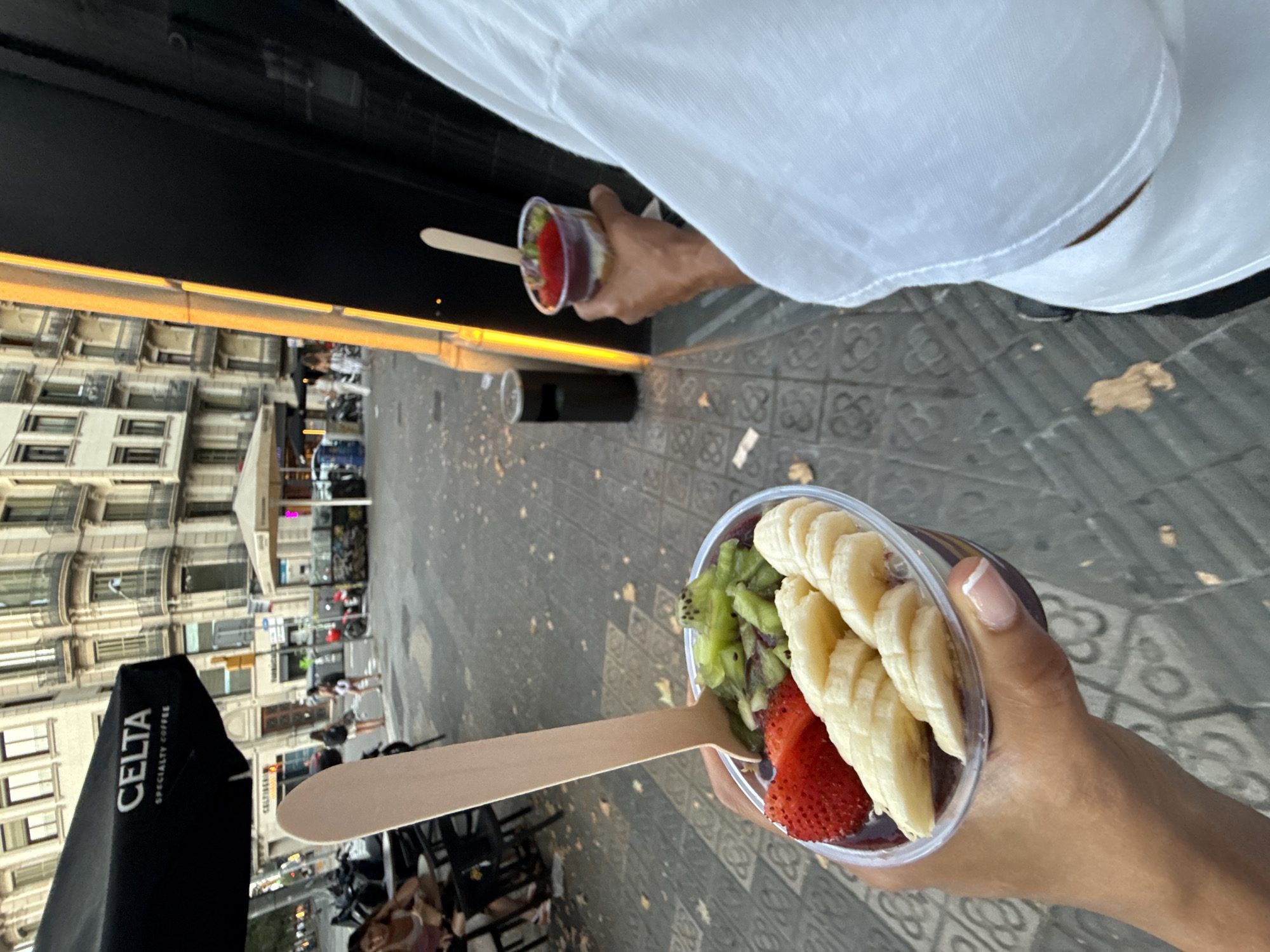The image captures a street scene in Barcelona, Spain, likely in an outdoor seating area of a cafe. In the foreground, a person is holding a clear plastic cup filled with an acai bowl topped with sliced strawberries, bananas, and kiwi. A wooden spoon is stuck into the bowl. To the right, another person, mostly out of frame and wearing a white t-shirt, holds a similar clear cup with a red fruit topping and a wooden spoon.
The background shows a cobblestone sidewalk with fallen leaves and a patterned tile border. Across the street, buildings with multiple stories and balconies are visible. A black umbrella with the word "CELTA" and "SPECIALTY COFFEE" printed on it stands prominently on the left. Outdoor cafe seating with chairs and tables are also present. In the middle ground, several people are walking and standing, and a few motorcycles are parked. A dark trash bin is visible on the right side of the sidewalk. The overall lighting suggests it is daytime.