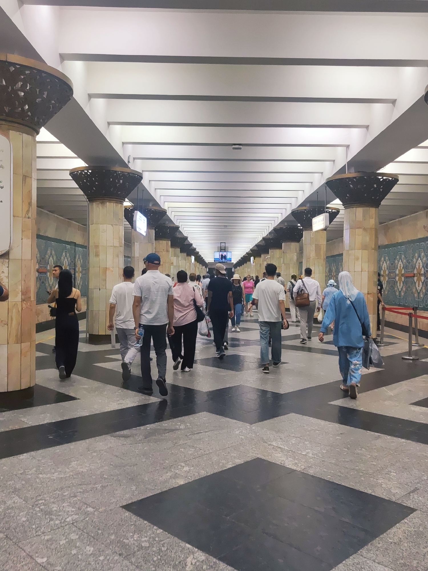 The image depicts an interior view of the Tashkent Metro, specifically at a station. The station features a long, wide corridor with a high, coffered ceiling supported by massive, ornately decorated pillars. The walls are lined with tiled panels featuring geometric patterns. A crowd of people, primarily dressed in casual attire, are walking in both directions along the corridor. The floor is paved with granite tiles in a pattern of black and grey squares and rectangles. The scene appears to be captured during the day, with artificial lighting illuminating the space. The overall impression is one of a busy public transit hub.