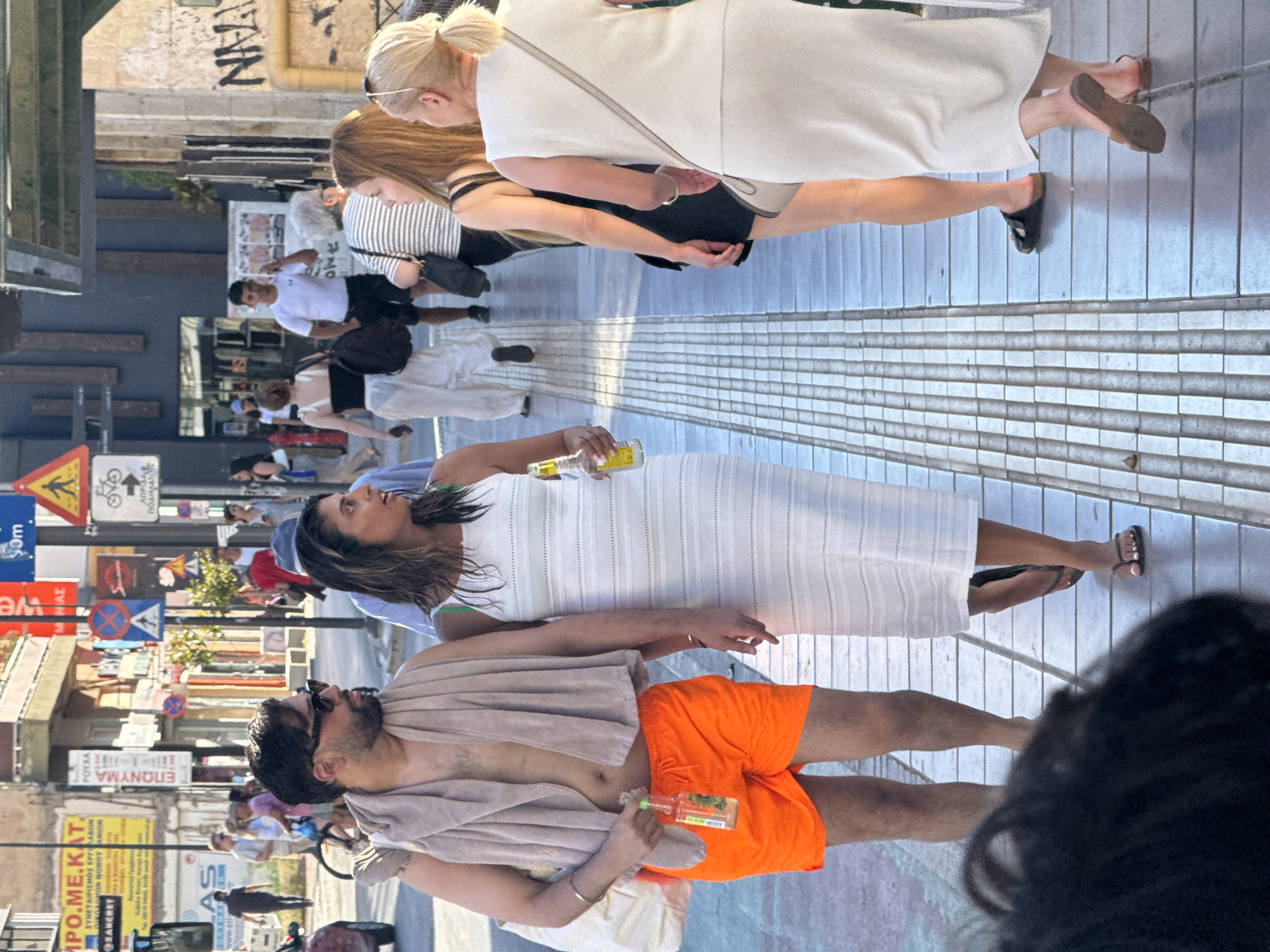 The image depicts a street scene in Chania, Greece. In the foreground, a man wearing bright orange shorts and a towel draped over his shoulders is walking next to a woman in a white dress. The man is holding a bottle of beverage, and the woman is also holding a bottle. They appear to be walking on a tiled sidewalk with a tactile paving strip. In the background, other pedestrians are visible, along with buildings, street signs, and a bus. The atmosphere suggests a sunny day, possibly during tourist season, given the casual attire of the people. The architecture and signage hint at a Mediterranean setting.