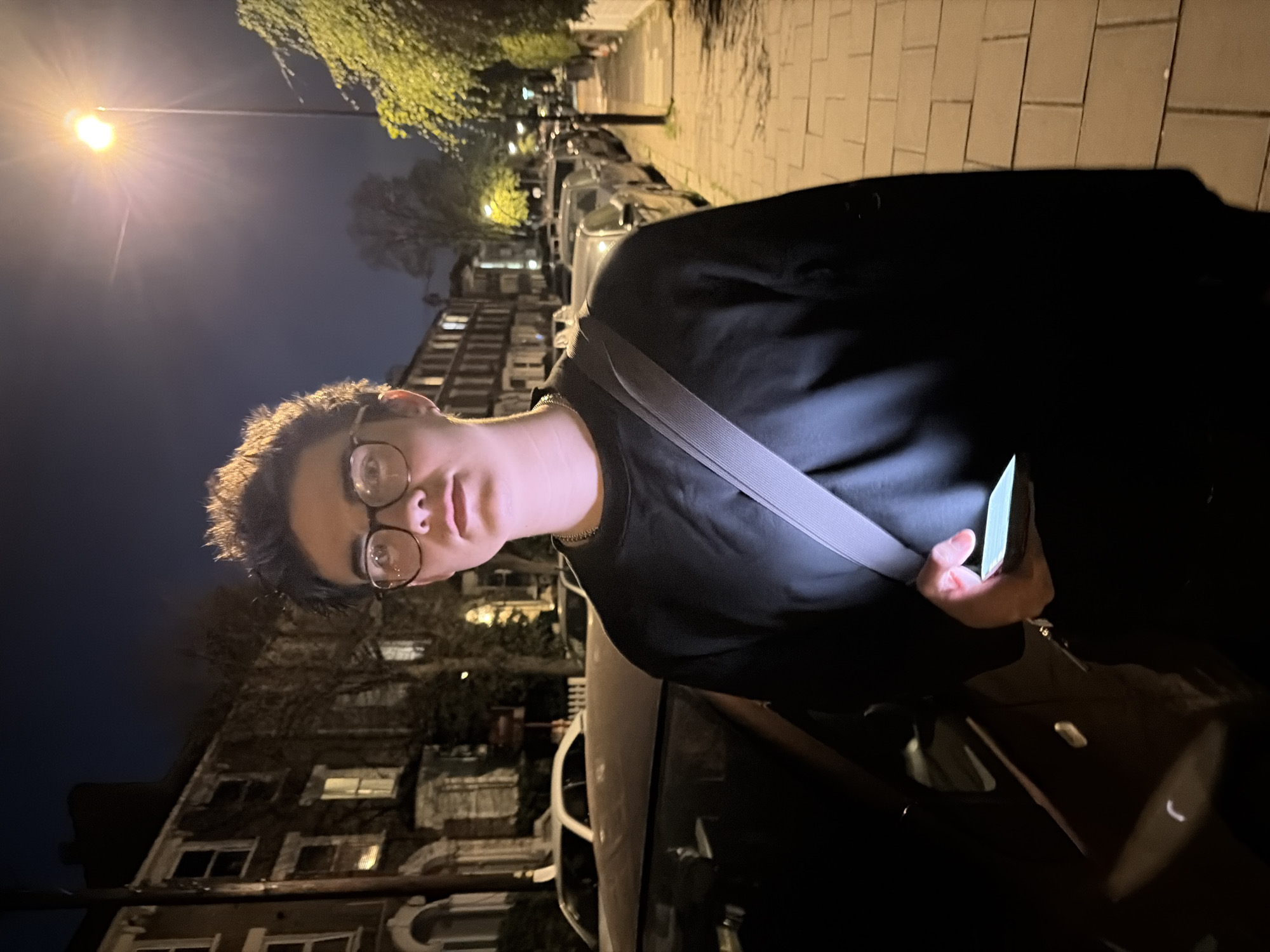 A young man with short brown hair and glasses is the primary subject, standing on a sidewalk at night in London, United Kingdom. He is wearing a black sweater and a grey strap across his chest, possibly for a bag. He is holding a smartphone in his right hand.
Behind him are rows of parked cars along the street. Across the street, there are multi-story brick buildings, typical of London residential architecture, with visible windows and architectural details. Street lamps are illuminating the scene. The sidewalk appears to be paved with rectangular slabs. The sky is dark, indicating it's either late evening or night. There is sparse foliage visible on trees bordering the buildings.