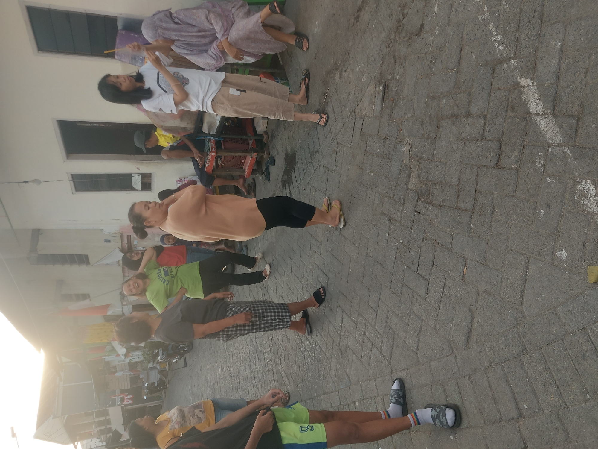 In an outdoor urban setting in Makassar, Indonesia, a group of people are gathered on a cobblestone street lined with buildings. The lighting suggests it is late afternoon or early evening, with the sun casting a warm glow from the left side of the frame.
Several individuals are standing and interacting. A woman in a green t-shirt with text "CALIGARAI WA.ART" is looking to her right with an open mouth, appearing to be speaking or laughing. Next to her, a woman in a dark t-shirt and plaid shorts is standing with her arms crossed. To the left, a woman in a yellow t-shirt is facing away from the camera. In the foreground, a person wearing bright green shorts and white socks with colorful stripes is partially visible, facing left.
Further to the right, a woman in a white t-shirt and tan pants is holding a thin stick or candle upwards, seemingly presenting it to another woman who is seated and partially covered by a purple headscarf and draped clothing. This seated woman is looking towards the woman with the stick. Behind them, a man in a yellow shirt and a dark cap is seated on a red crate.
The environment includes buildings with visible windows and doors, some adorned with flags. A motorcycle is parked in the background. The ground is paved with interlocking cobblestones, and there are some scattered debris and white markings on the pavement. The overall scene depicts a casual gathering of people in a residential or neighborhood street.