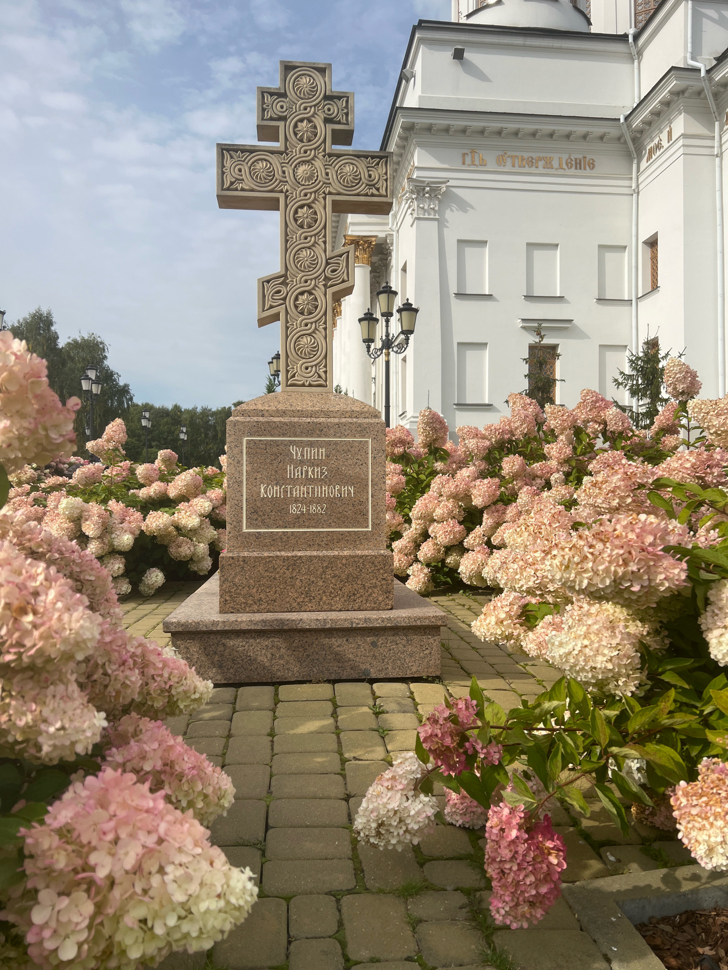 The image depicts a large, ornate stone cross standing on a granite pedestal in front of a white building. The cross is decorated with intricate carvings. The pedestal has an inscription in Russian which translates to "Chupin Narkiz Konstantinovna 1824-1882". The area around the pedestal is paved with interlocking bricks and surrounded by lush bushes with pink and white hydrangeas. In the background, the white building appears to be a church or cathedral, with architectural details suggesting an Orthodox Christian style. The sky is partly cloudy. This monument is located in the Yekaterinburg City Historical Museum, specifically at the Church of All Saints.