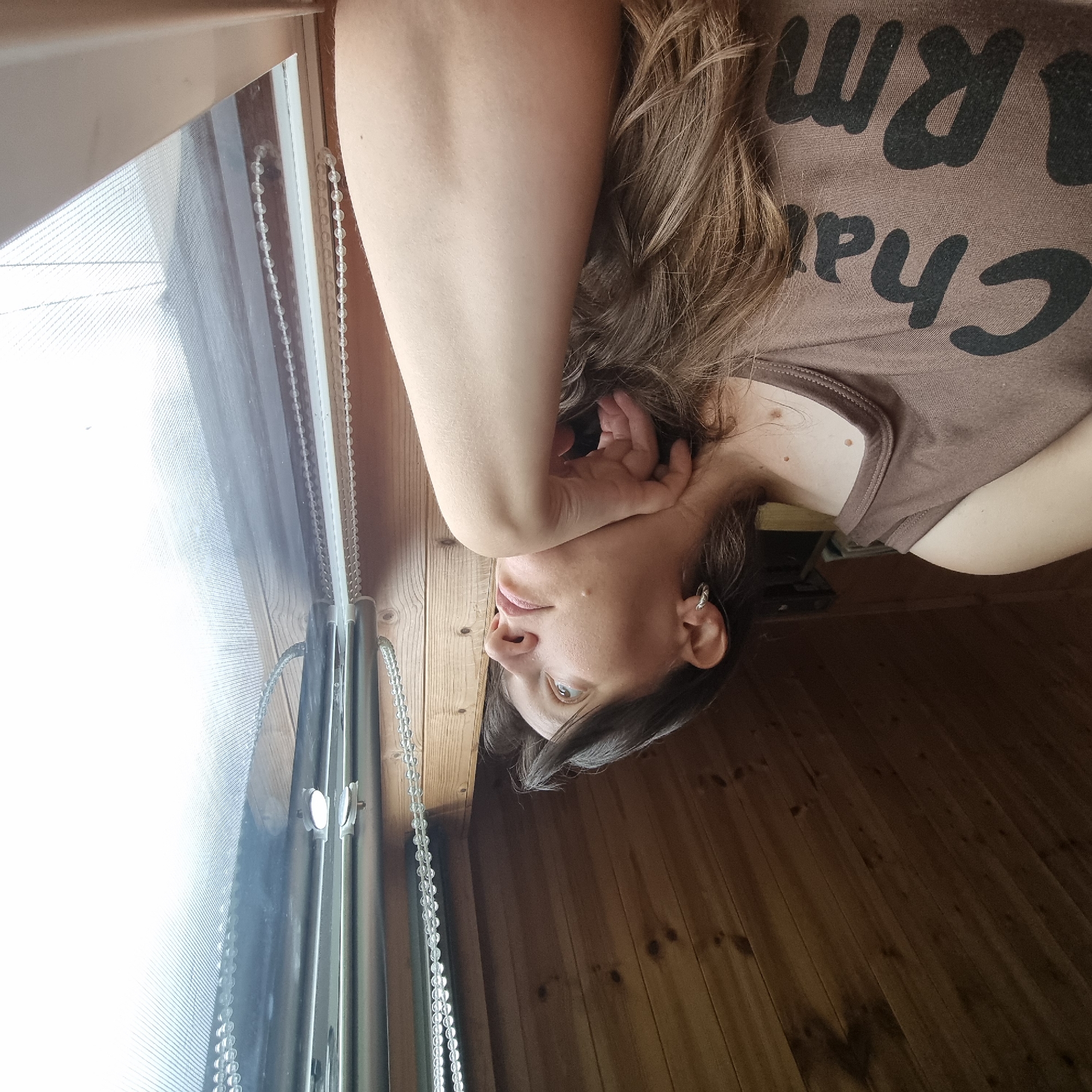 A woman with long, dark brown hair, wearing a brown tank top with text on it, is resting her head on her hand as she looks out a window. The interior appears to be a wooden room, with a sloped wooden ceiling visible at the top of the frame. A beaded blind cord hangs to the right of the woman, near the window. The scene is lit by natural light coming from the window. The image was taken in Dunnae-myeon, South Korea.