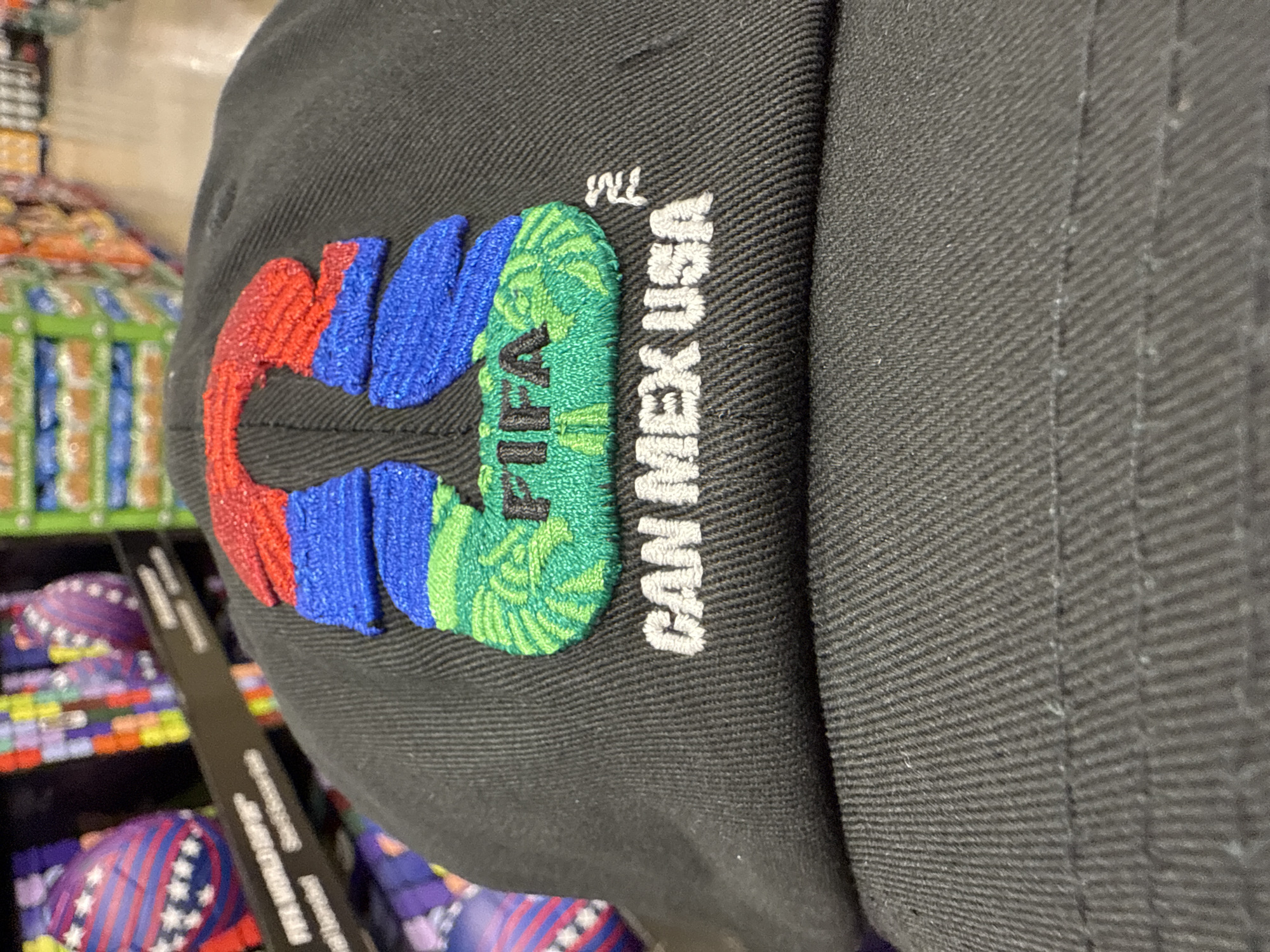 The image shows a close-up of a black baseball cap. Embroidered on the cap is the logo "CAN" in red, blue, and green letters, followed by "MEX USA" in white letters. The green letter also has "FIFA" written in black. The cap appears to be in a store in Bolingbrook, United States of America, as there are shelves of merchandise visible in the background, including items with stars and stripes patterns.