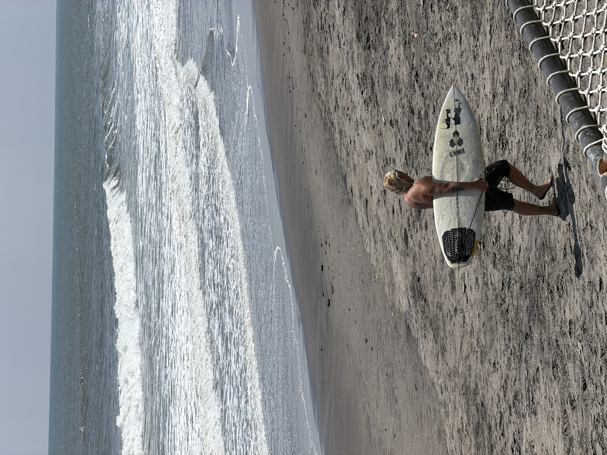 A male individual with blonde hair, wearing dark shorts, walks barefoot across a dark sand beach in Belalang, Indonesia. He carries a white surfboard, featuring a black traction pad and "Al Merrick" branding, under his right arm. Tattoos are visible on his left arm. The background depicts the ocean with multiple white-capped waves breaking towards the shore, under a pale grey sky. The dark sandy foreground contains scattered pebbles and visible footprints. A dark cylindrical barrier with white netting is partially visible in the lower right corner.