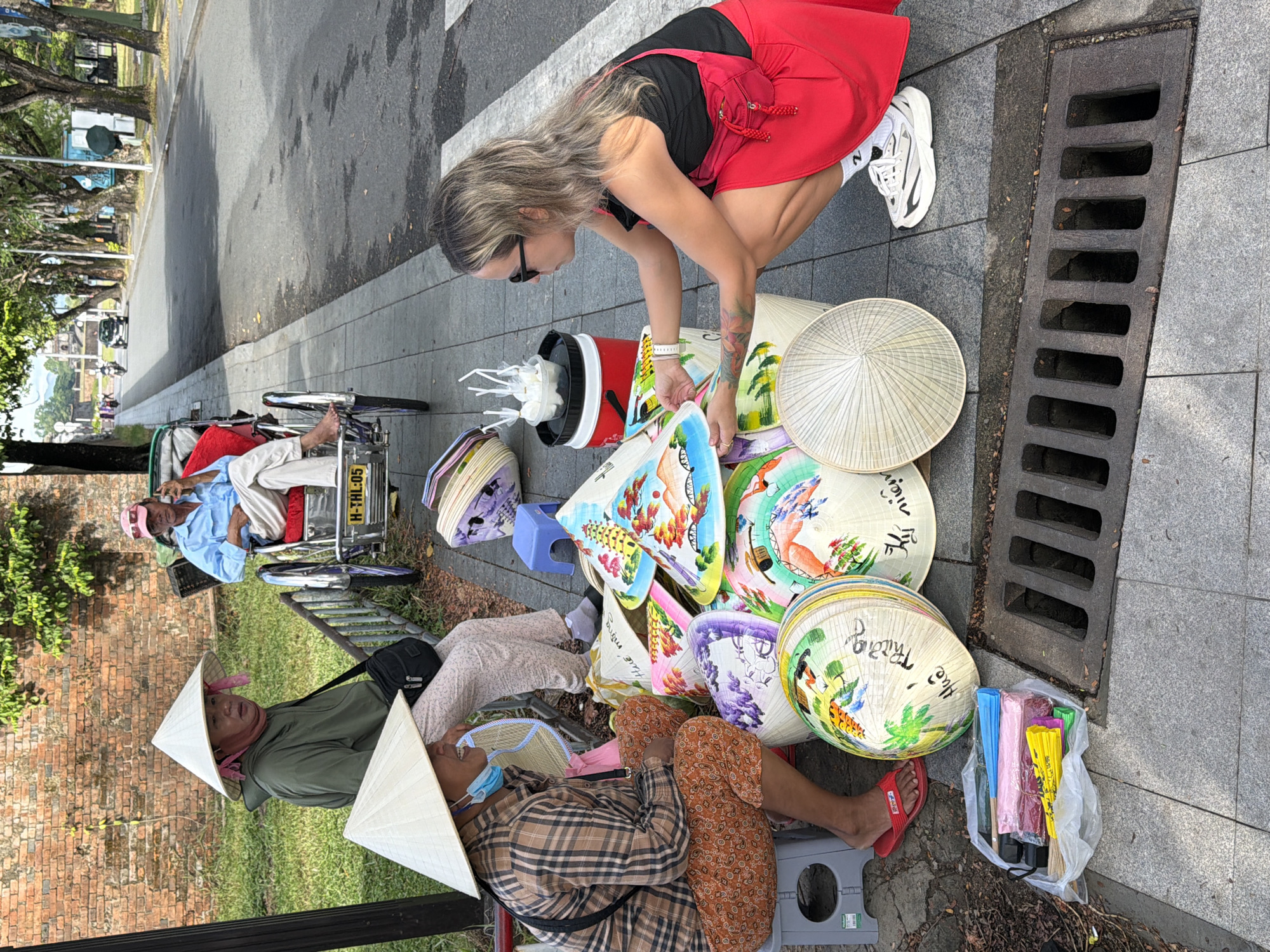 In Phú Xuân Ward, Vietnam, a woman wearing sunglasses and a red skirt squats to examine a pile of conical hats displayed on a sidewalk. She is wearing a black tank top and white athletic shoes. Next to her, a woman in a plaid shirt and conical hat is seated on a small stool, looking away from the camera. Behind them, another woman in a green jacket and conical hat is seated in a pedicab. A man in a blue shirt sits in the pedicab, holding a mobile phone to his ear. The hats are decorated with colorful paintings, and some have Vietnamese writing on them. In the foreground, a plastic bag contains folded umbrellas in various colors. The sidewalk is paved with gray tiles, and a metal grate for drainage is visible. Trees line the street behind the pedicab, and a brick wall is present on the left side of the frame. The scene appears to be a street market or a roadside stall.