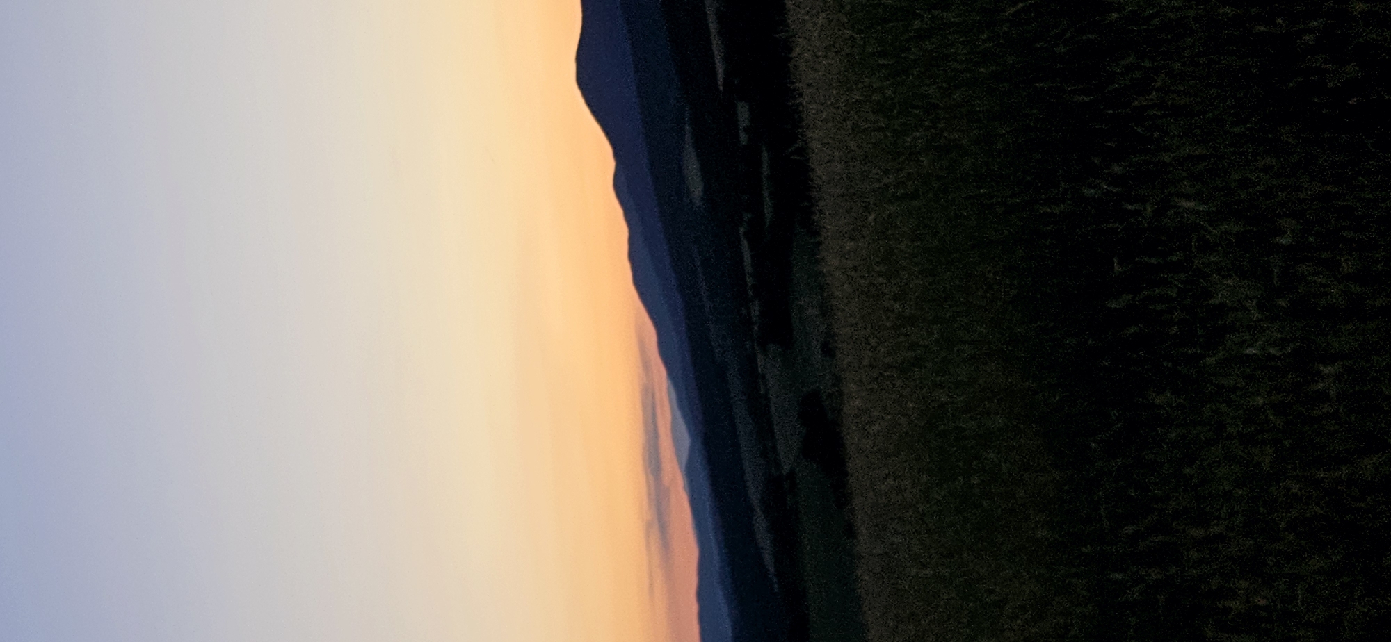 The image shows a rural landscape at dusk. In the foreground, tall grass is visible, appearing dark and silhouetted against the fading light. Behind the grass, rolling hills and mountains are visible, also largely in shadow, with some appearing as darker shapes against the sky. The sky itself is a gradient of colors, transitioning from a pale blue at the top to a warm orange and pink near the horizon, indicating the sun has recently set or is about to rise. The overall impression is one of natural beauty and tranquility. The image is a vertical composition.