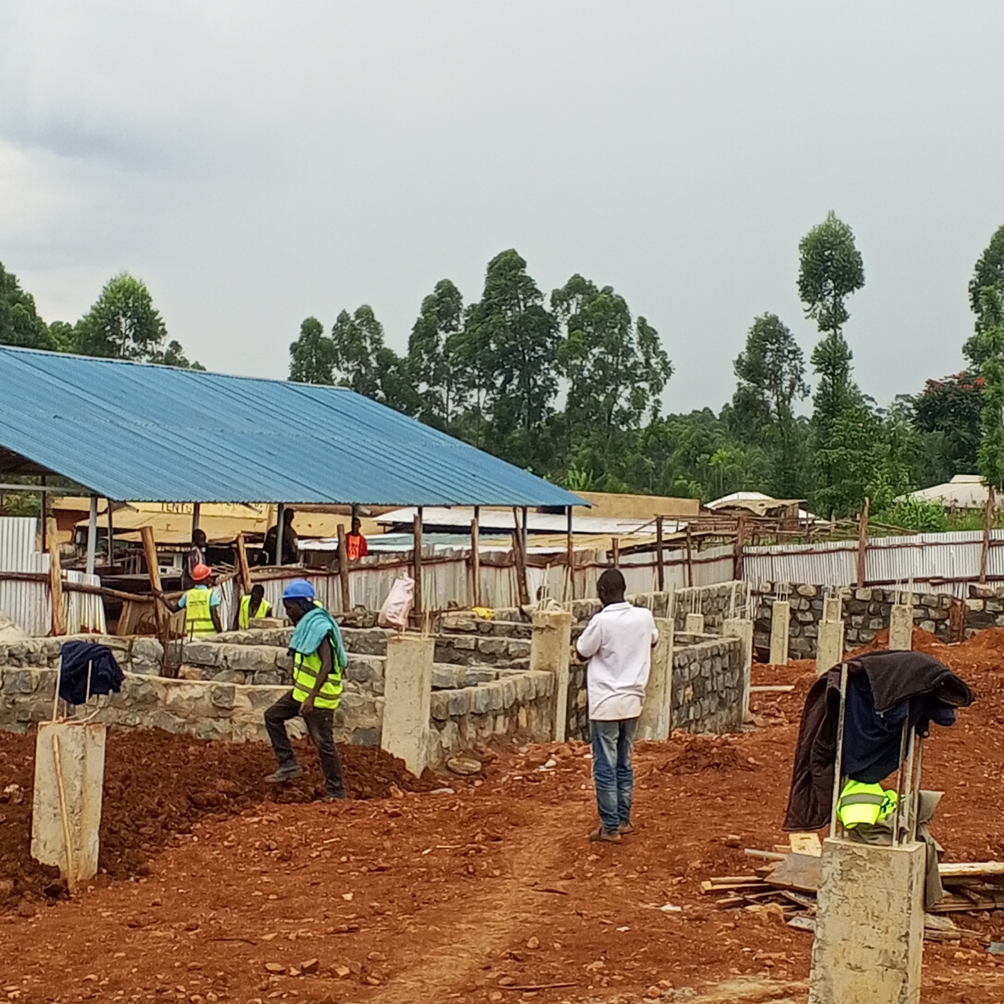 This media file captures an active construction site in Borabu Chitago ward, Kenya, under an overcast sky. The ground is predominantly exposed reddish-brown soil, characteristic of the region, indicating ongoing earthworks.
In the foreground and mid-ground, the foundations of a new building or complex are visible. These foundations consist of rough-hewn stone blocks mortared together, forming low walls, interspersed with concrete pillars. Several workers are present on the site. On the left foreground, a worker wearing a blue hard hat, a yellow safety vest, and dark trousers walks across a mound of dirt. In the mid-ground to the right, another person in a white t-shirt and blue jeans walks away from the camera.
Towards the background, a large structure with a bright blue corrugated metal roof stands, possibly a temporary shelter, storage area, or an existing building being expanded. Beneath this roof, several other workers are visible, some also wearing yellow safety vests. Corrugated metal sheets are used as fencing or temporary partitions around parts of the site. In the distance, a dense line of green trees suggests a verdant, possibly rural or semi-rural, setting.
Notable details include construction materials like wooden planks scattered on the ground. A dark jacket, a yellow safety vest, and a blue towel-like item are hanging on one of the concrete pillars in the right foreground. Faint, partially obscured text, possibly "TENIS" or "TENTG," can be seen on a banner or sign attached to the blue-roofed structure in the background. The scene depicts a busy construction environment in broad daylight.
