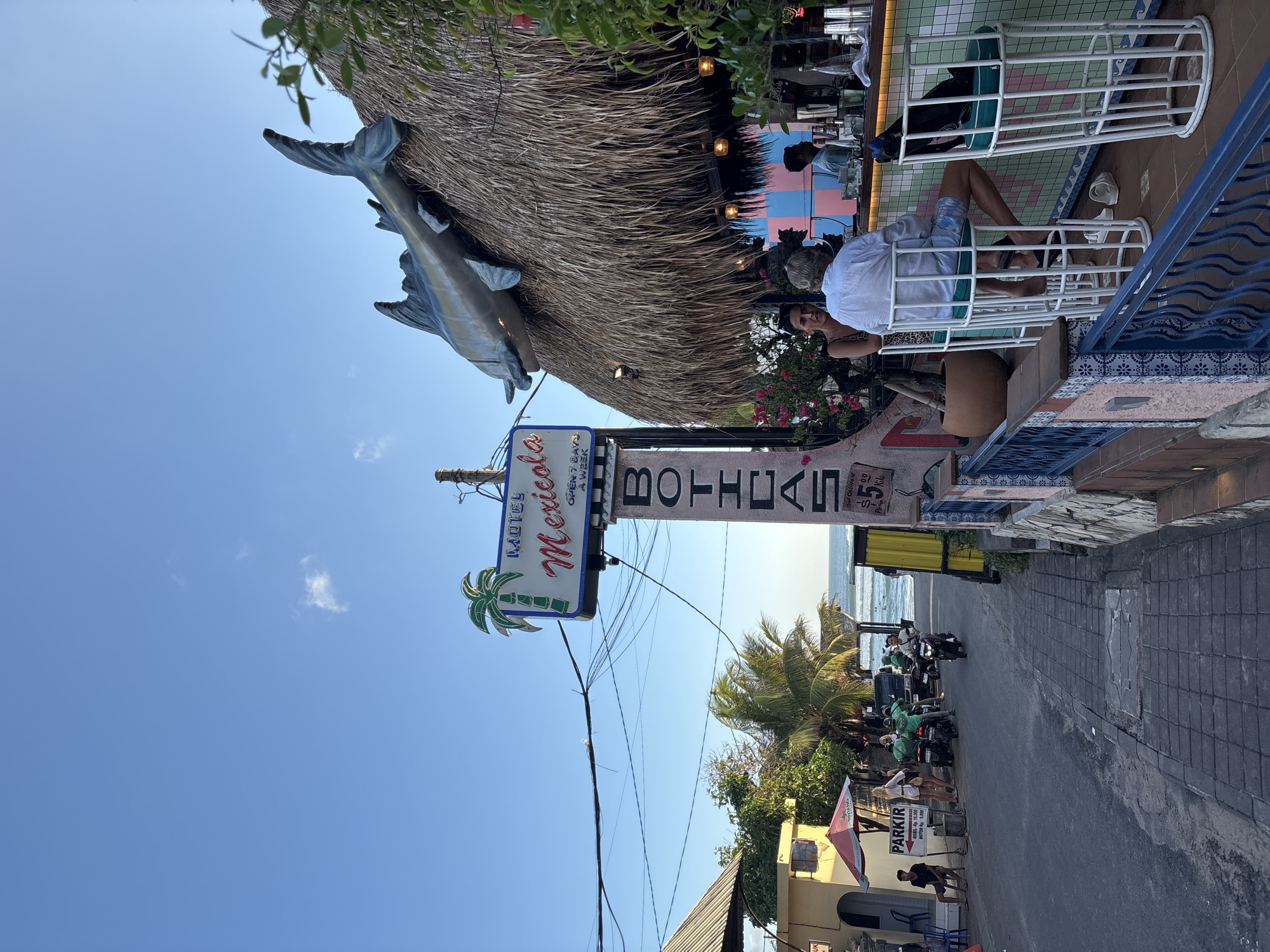 Here is a description of the image:
The image depicts the Motel Mexicola in Canggu, Indonesia. The motel features a thatched roof with a decorative fish mounted on it. Patrons are seated in white chairs on a patio, with a bar visible in the background. A sign reading "Motel Mexicola" is displayed prominently. Below it, a sign reading "Boticas" is affixed. The street scene shows motorcycles and pedestrians. The ocean is visible in the distance. The sky is blue.