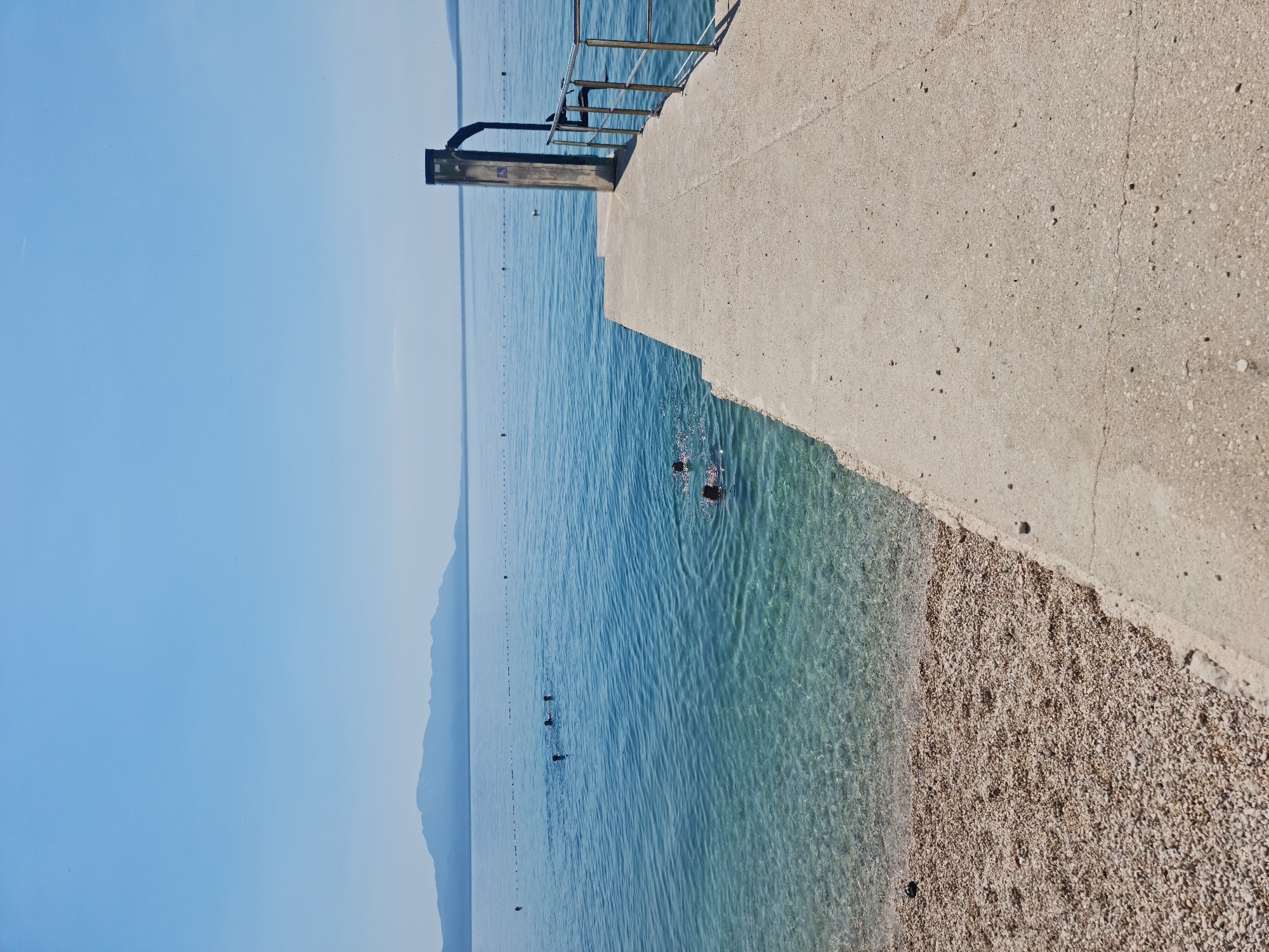 The image captures a sunny day at a beach in Općina Gradac, Croatia. A concrete pier extends into the clear, blue water from a pebbly shore. Several people are visible swimming in the sea, their heads bobbing above the surface. The water is transparent, revealing the rocky seabed. In the distance, a mountain range forms a hazy silhouette against the bright blue sky. The pier is equipped with a metal railing and a tall, metallic structure, possibly a shower or a lifeguard station. The overall scene conveys a sense of leisure and a typical summer day at the Croatian coast.