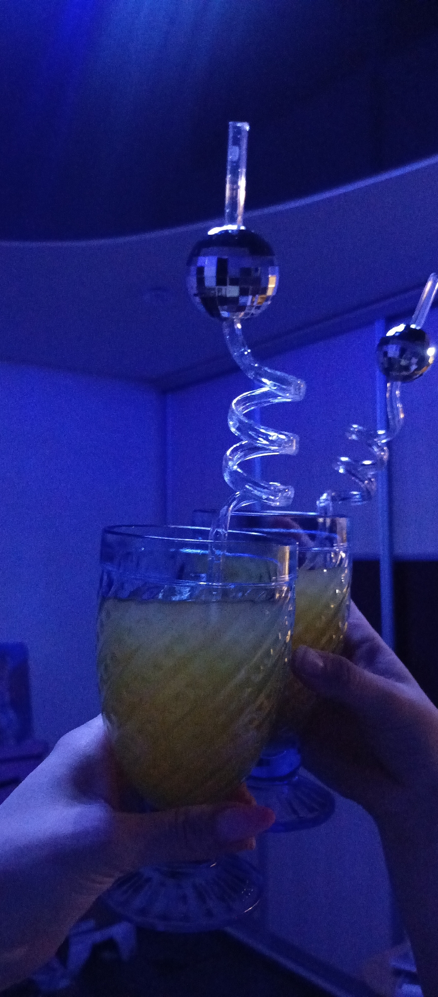 Here is a detailed description of the image:
**Scene:** The image depicts two stemmed glasses filled with a yellow liquid, held up in what appears to be a celebratory toast.
**People:** Two hands are visible holding the glasses. The hands are light-skinned.
**Subjects:** The main subjects are the two glasses and their contents. The glasses are decorated with a textured pattern. The yellow liquid inside is unclear if it is a cocktail, juice, or another beverage.
**Objects:** Unusual spiral straws with disco ball on top.
**Actions:** A celebratory toast or the act of raising glasses.
**Interactions:** Two people hold the glasses.
**Environment:** The background suggests an indoor setting, possibly a bar or club, with blue-toned lighting. The room appears relatively simple in terms of décor, with a wall and corner visible in the background.
**Location:** The city of Unknown, Unknown.