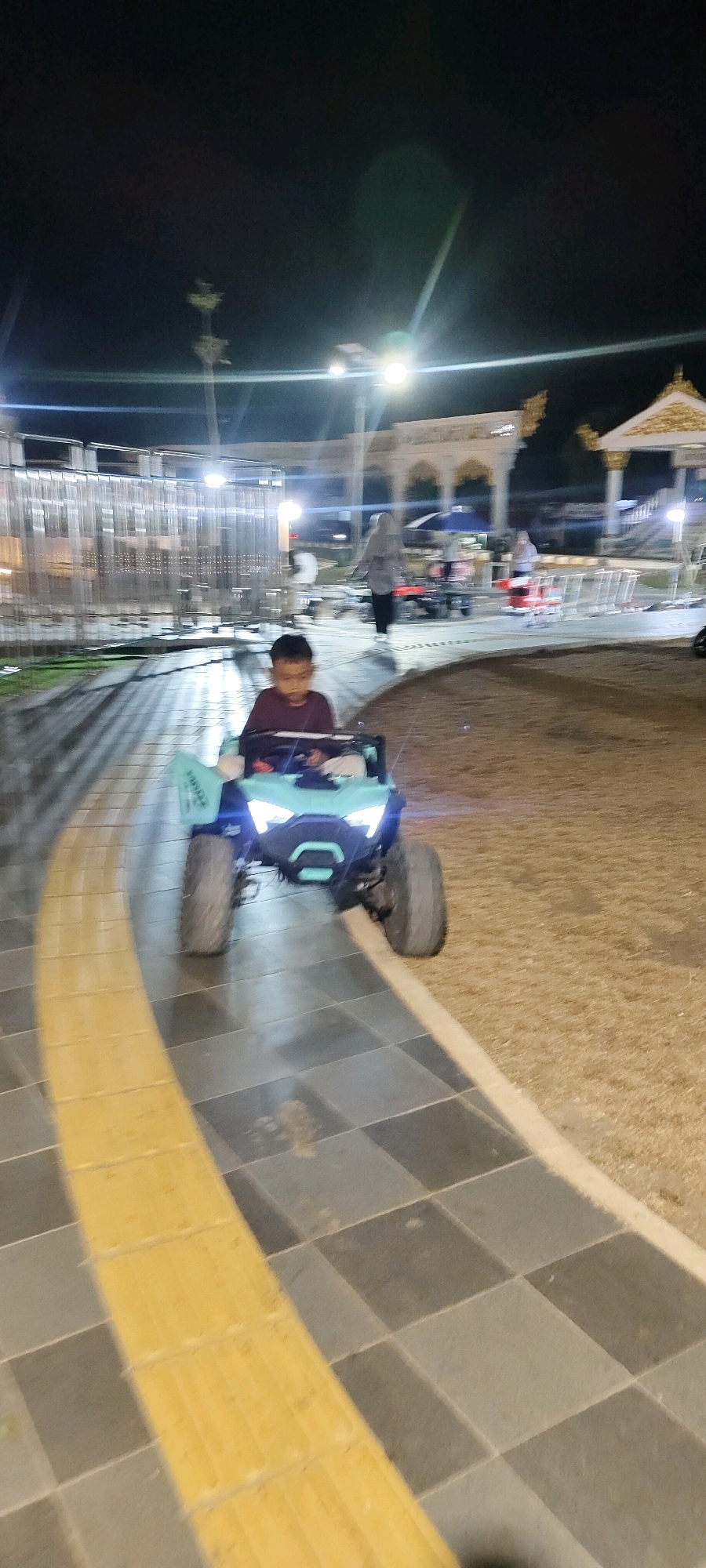 In Martapura, Indonesia, a young boy is pictured riding a child-sized electric toy car at night. The car, a bright teal color, has illuminated headlights and large tires. The boy, wearing a dark red shirt, is seated in the driver's position. The scene takes place on a paved walkway with contrasting yellow and dark gray tiles. In the background, there are blurred figures of people, a street light, and architectural structures, suggesting a public space like a park or recreational area. The overall impression is one of nighttime entertainment.