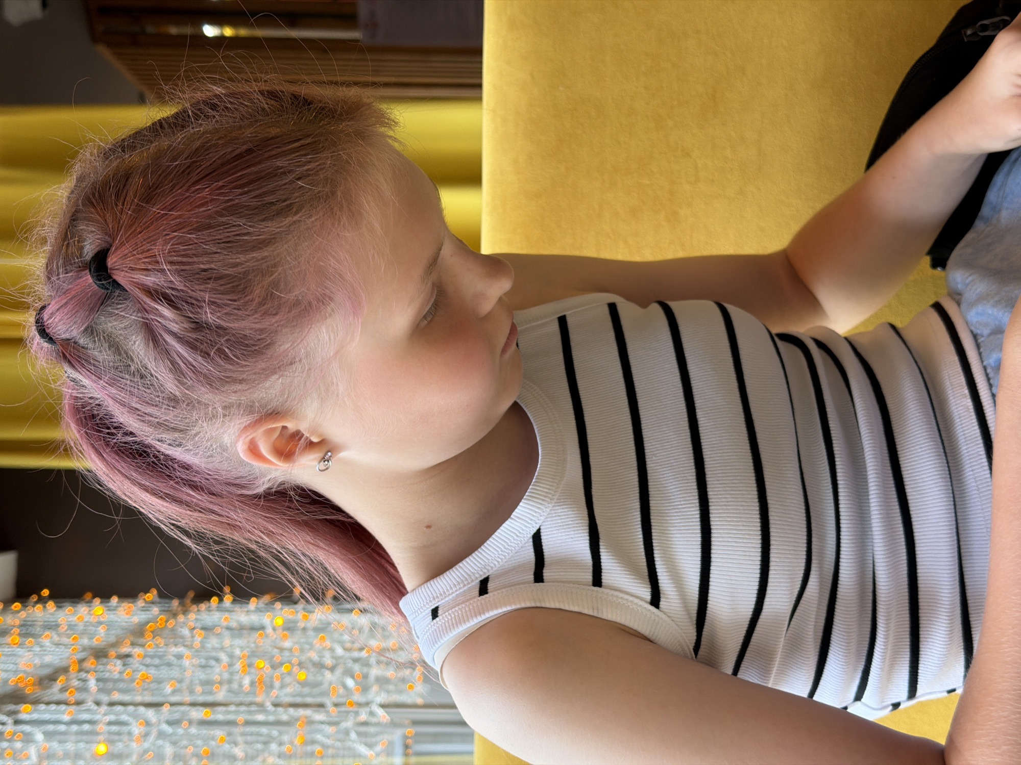 The media file shows a close-up, rotated view of a young girl with pink-tinged hair, wearing a white ribbed tank top with black stripes. She is lying down, her head propped up on a yellow surface, possibly a couch or cushion. Her eyes are closed, and her face is turned to the side, suggesting she is resting or sleeping. A small, silver hoop earring is visible in her left ear.
In the foreground, out of focus, are numerous small, glowing orange lights, suggesting a decorative string of lights. The background is largely occupied by the yellow surface, with some dark wooden elements and a blurred area with more lights. The lighting in the image is warm and soft, indicating it might be evening or an indoor setting with artificial light. The overall mood is peaceful and relaxed. The image appears to have been taken indoors, in Chaikovsky, Russia, based on the provided location context. There is no visible text in the image.
