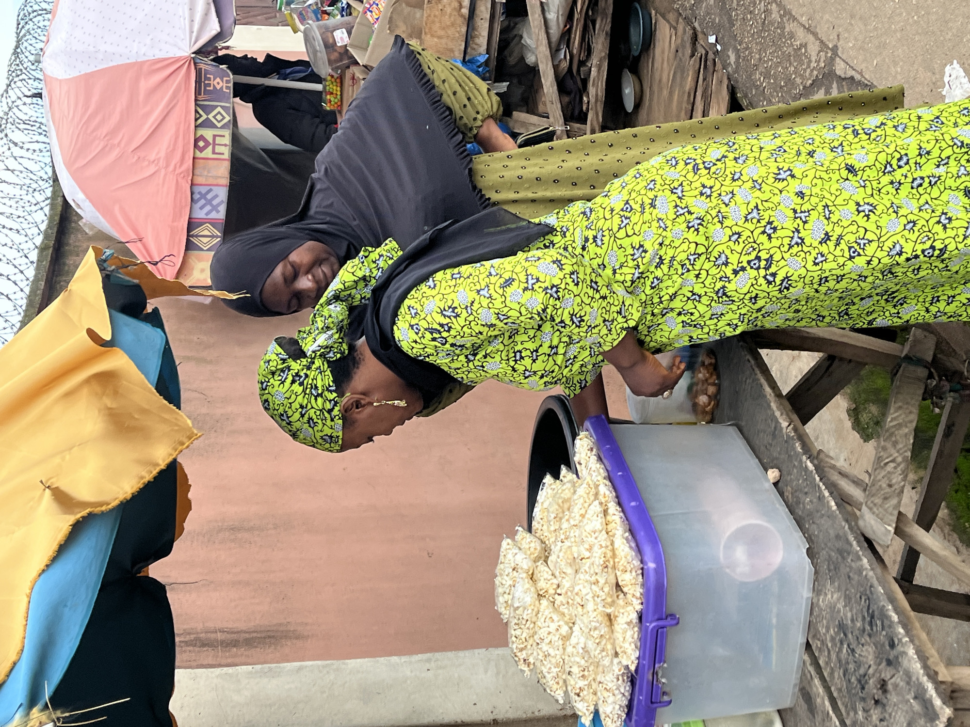 Two women are standing at a market stall under a canopy in Jos, Nigeria. The woman in the foreground is wearing a bright green dress with a black and white floral pattern and a black headscarf. She is reaching into a clear plastic container filled with bags of popcorn. The woman behind her is wearing a dark hijab and a dark green dress with a polka dot pattern. She is looking down at the popcorn. The stall is made of wood and covered by a yellow and blue umbrella. There are other market stalls and supplies visible in the background. The ground is dirt and concrete.