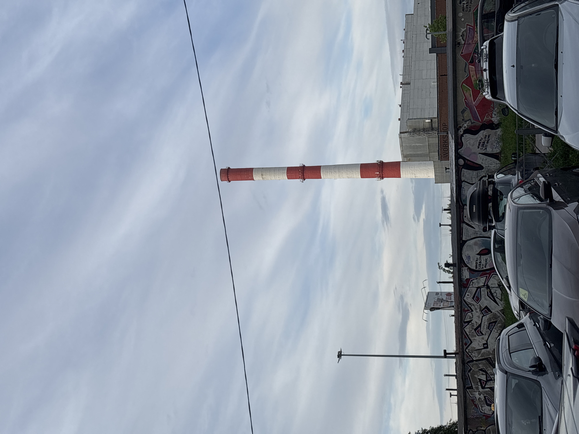 This image, taken on an overcast day, captures an industrial or urban scene primarily featuring a tall, red and white striped industrial chimney. The chimney stands prominently in the mid-ground on the left, extending vertically towards a pale, cloudy sky. A dark utility cable runs diagonally across the upper left portion of the frame.
To the right of the chimney, several parked cars are visible, their dark and silver bodies reflecting the soft light. Behind these cars, a substantial concrete wall is covered in extensive graffiti. Above this wall, a multi-story building, likely part of an industrial complex, can be seen, featuring a series of rectangular structures or windows along its top.
Notable details include a light pole with a luminaire head positioned in the mid-ground to the right of the chimney. The sky is largely covered with thin, wispy clouds, indicating daytime but not direct sunlight. The graffiti on the wall displays various styles and colors, including stylized letters in red and black, and a more detailed piece depicting a face-like figure with text.
Visible text elements include "WORKSHOP" written on a section of the wall or building. Among the graffiti, phrases such as "MAMU KTO ZABRAL TSU 27.09.1977 14:00" and "AVTUINO" are discernable, possibly in a Cyrillic script, suggesting a specific cultural or personal message. No people are visible, and the scene appears static, focusing on the structures and vehicles within this urban-industrial landscape.