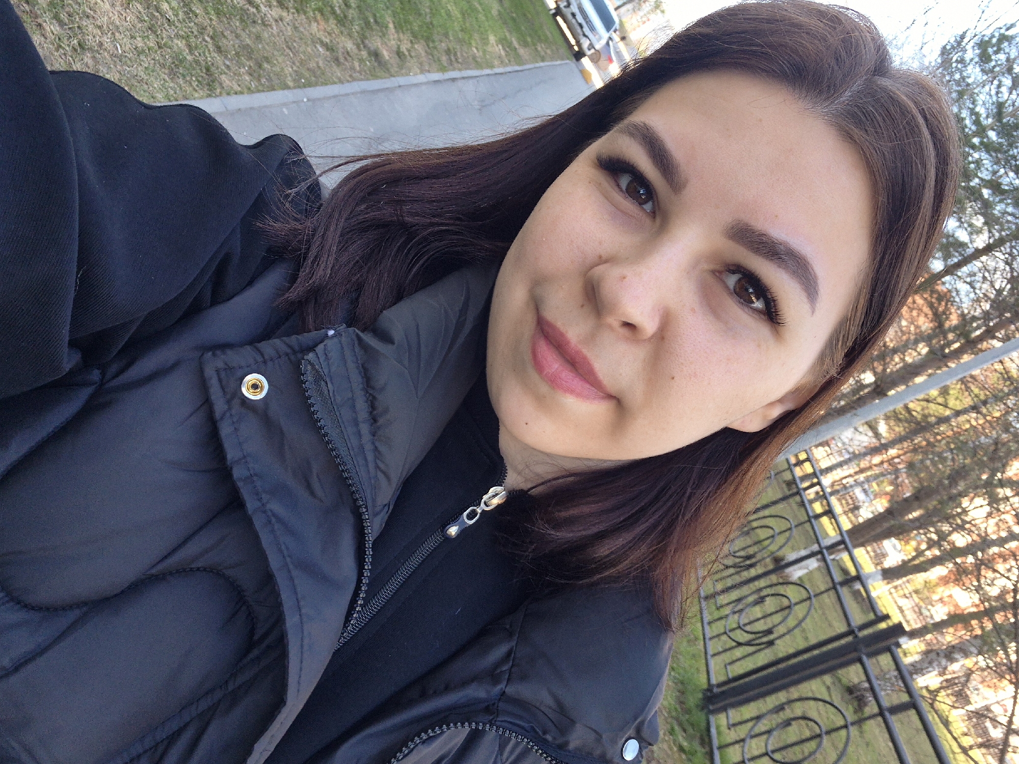 A selfie of a light-skinned woman with brown hair and defined eyebrows, wearing a black puffer jacket, taken outdoors. The background shows a sidewalk, grass, trees, and a metal fence. The location is possibly in Tobolsk, Russia.