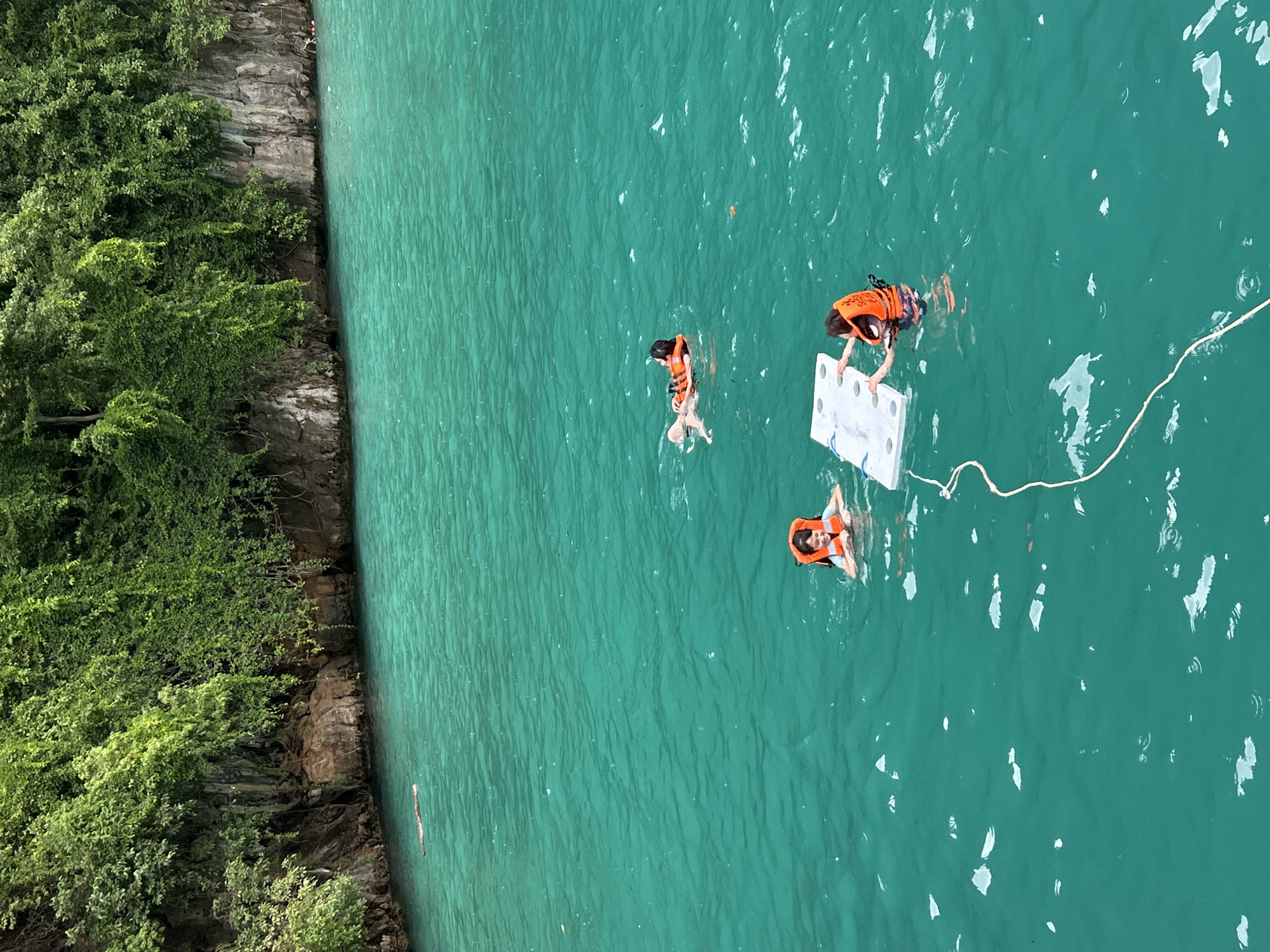 In Ao Nang, Thailand, three individuals are observed swimming and interacting with a floating raft in vibrant turquoise waters. The scene is framed by a lush green cliff face covered in dense vegetation, suggesting a tropical environment.
One person is floating on their back, wearing an orange life vest. Another individual is positioned near the raft, also wearing an orange life vest, and appears to be pushing or stabilizing it. A third person, wearing an orange life vest, is partially submerged in the water, looking towards the raft. The raft itself is white and rectangular, with a rope attached. Scattered white debris floats on the water's surface. The water is clear, with visible ripples and a slight wave pattern. The overall impression is one of recreational activity in a picturesque natural setting.