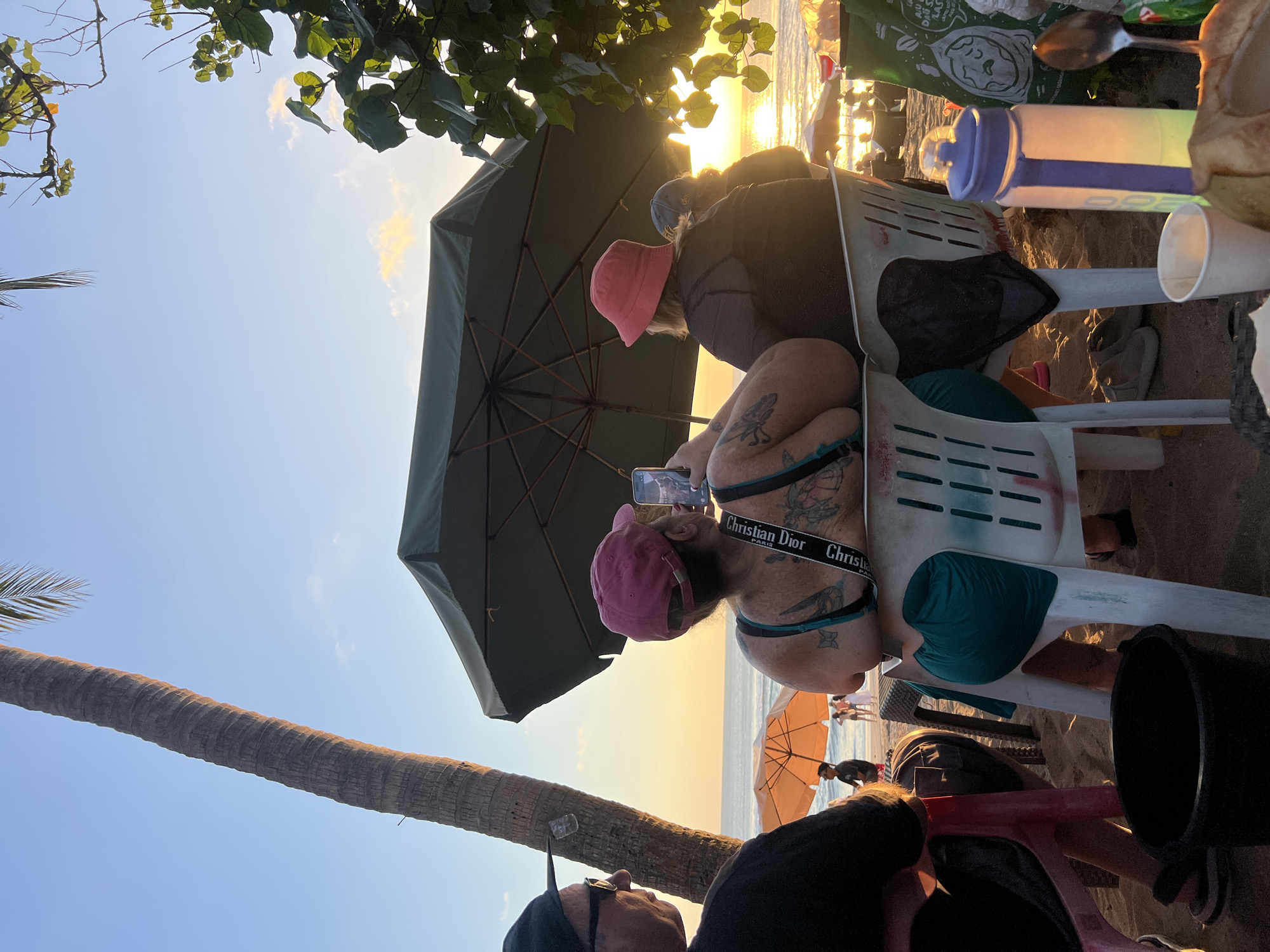 The image depicts a sunset scene on Kuta Beach, Indonesia. In the foreground, two individuals are seated on plastic chairs facing away from the camera, towards the ocean. The person on the left wears a dark t-shirt and a baseball cap, while the person on the right wears a pink bucket hat and a dark top, holding a smartphone. Both have tattoos visible. A large dark green umbrella is positioned behind them, casting a shadow. To the left, a palm tree trunk stands tall, with its fronds extending out of frame. In the middle ground, other beachgoers are visible, some seated under an orange umbrella. The ocean stretches to the horizon, reflecting the warm colors of the sunset. The sand is the primary ground cover. Various objects are present, including a black bucket in the lower left, a purple and white water bottle, a white paper cup, and a spoon on a table in the foreground, along with what appears to be a coconut. The overall atmosphere suggests a relaxed evening by the sea.