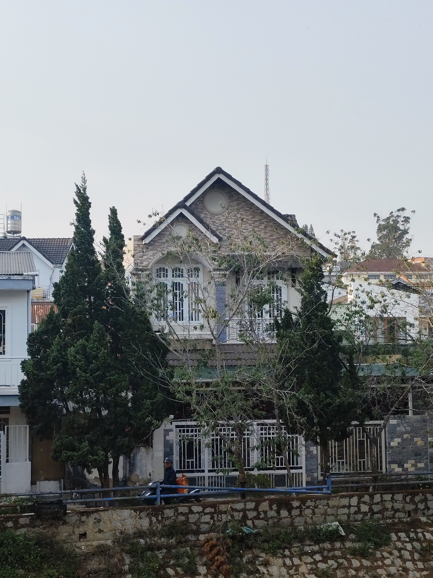 The image depicts a two-story house in Da Lat, Vietnam. The house has a stone facade on the second story and a gray roof. There are tall, dark green coniferous trees in front of the house, partially obscuring the view. A bare tree with thin branches is also visible. In front of the house is a metal fence, and behind it, a person on a scooter is traveling along a road. The foreground consists of a low stone wall and some vegetation. Other houses are visible in the background, suggesting an urban or suburban setting. A tall antenna is also visible. The sky is overcast and gray.