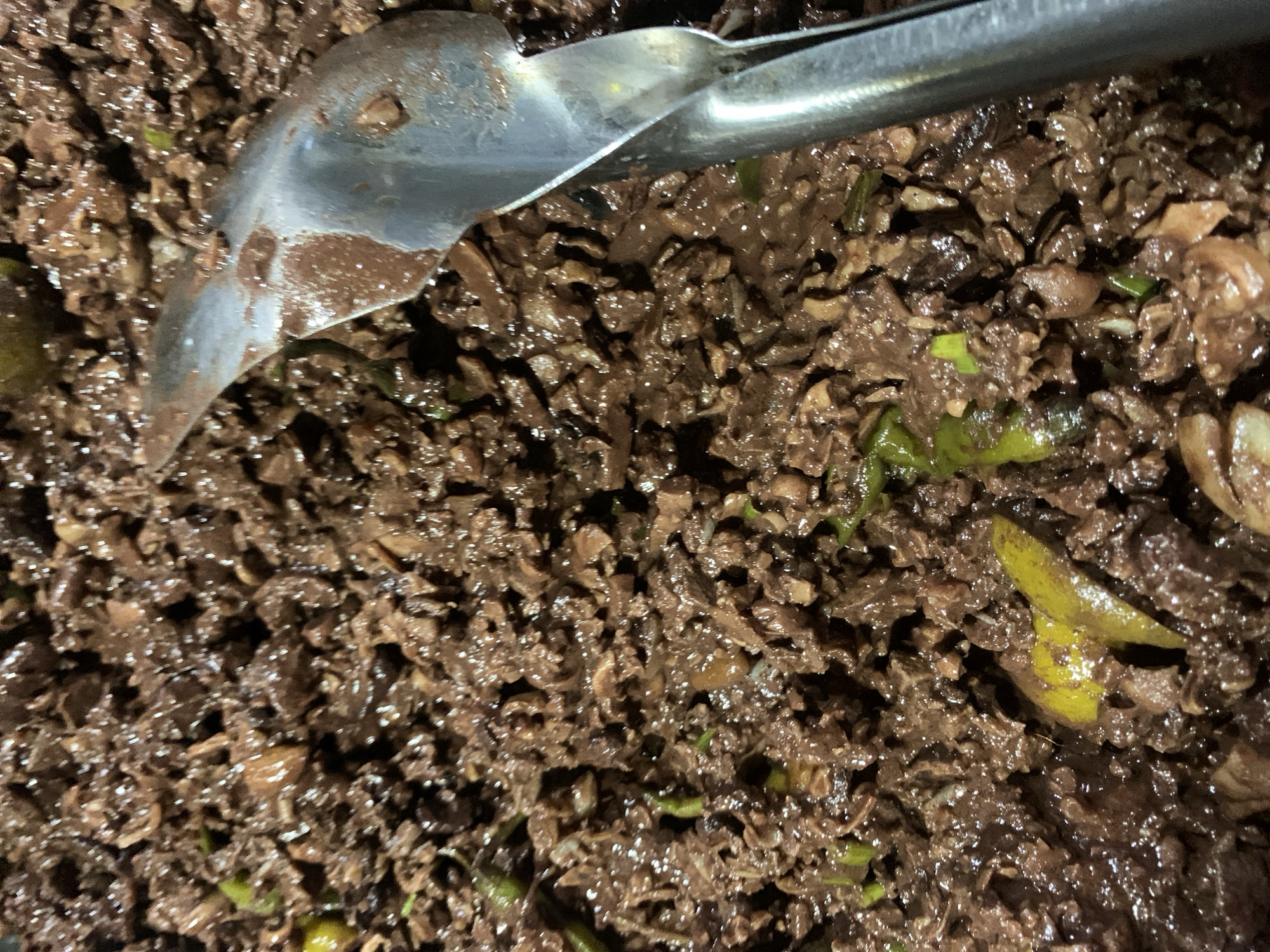 This image shows a close-up, overhead view of a dark, finely chopped dish being served with a metal spoon in Kiblawan, Philippines. The dish appears to be a savory mixture, possibly meat or blood-based, with visible small green pieces, likely chili or herbs, and some yellow slivers, possibly ginger or lime. The texture is moist and slightly clumpy, with a glossy sheen. The spoon, made of polished metal, is positioned in the upper right quadrant of the image, with its edge resting in the food, suggesting it is in the process of being served or is already in use. The lighting is dim, creating shadows and highlighting the textures of the food. The overall impression is of a hearty, possibly traditional, dish being prepared or consumed in a domestic or communal setting in Kiblawan.