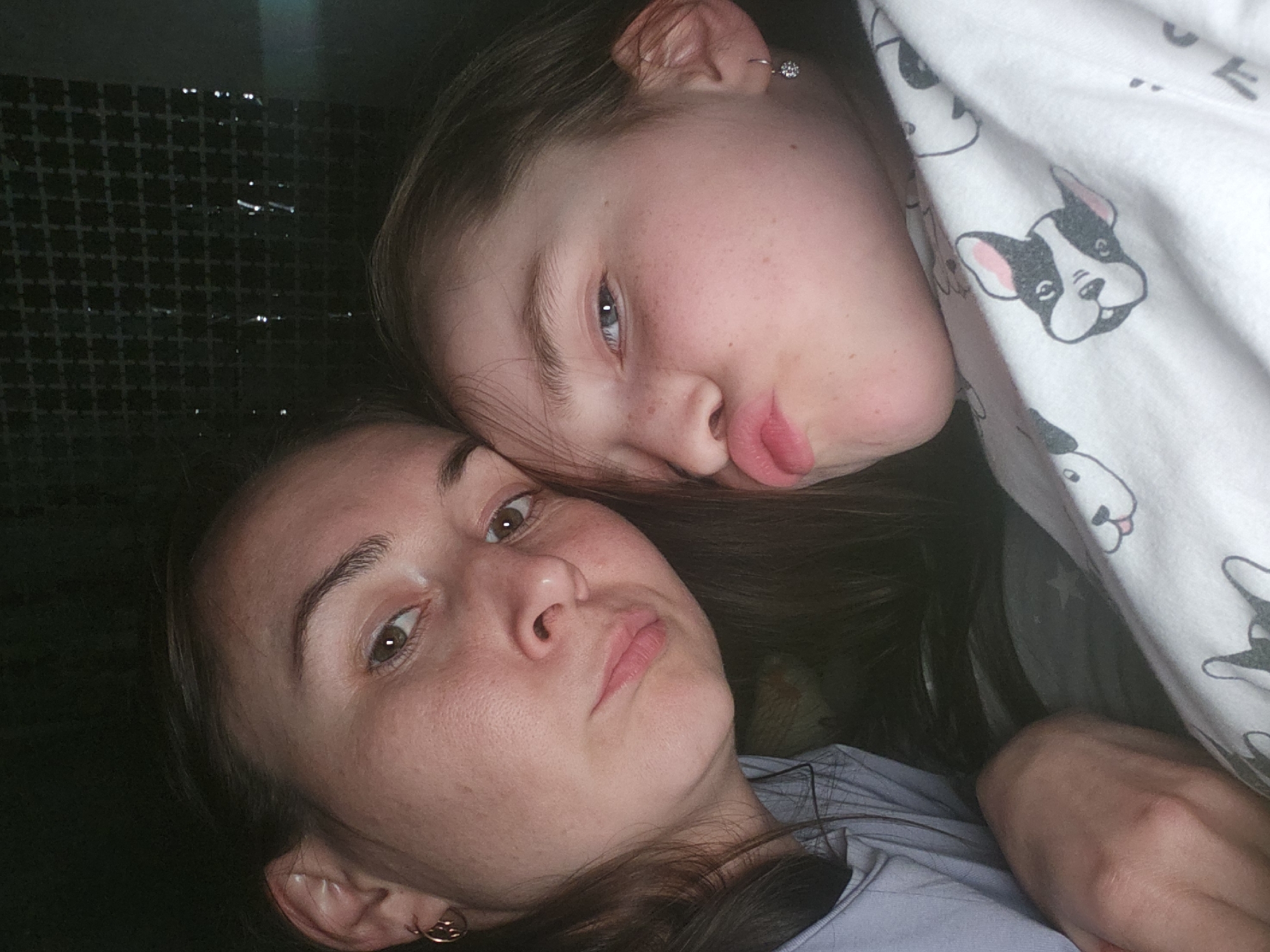 Two individuals, likely a mother and daughter, are positioned close together and looking at the camera. The younger individual is resting their head on the older individual. The younger individual is partially covered by a white blanket with cartoon dogs printed on it. The older individual is wearing a light blue shirt. Their facial expressions are similar, both with pursed lips. The background shows a wall with a grid pattern on the left side. The location is Krasnogorskoye, Russia.
