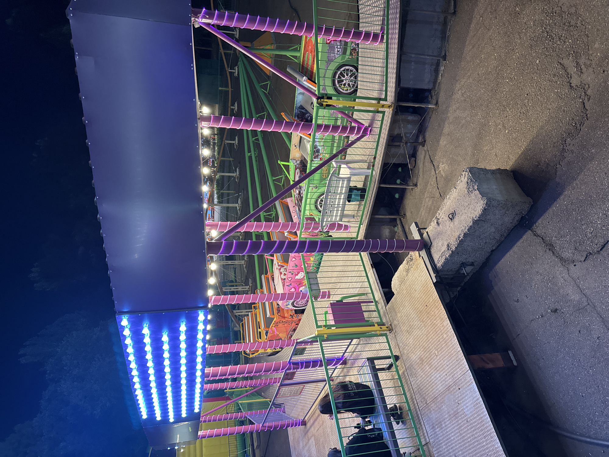 This is a nighttime exterior shot of an amusement park ride, likely a roller coaster, in the city of Oskemen, Kazakhstan. The ride features bright blue LED lights along the top edge of a purple awning and purple spiral lights wrapped around vertical poles. Several colorful, car-shaped ride cars are visible on the track, including one orange car with a Hello Kitty design and a green car with Star Wars-themed graphics. Two individuals are seated on a bench on a metal platform, facing away from the camera. The ground appears to be asphalt, and a large concrete block is present in the foreground. The overall environment suggests a festive, outdoor entertainment setting.