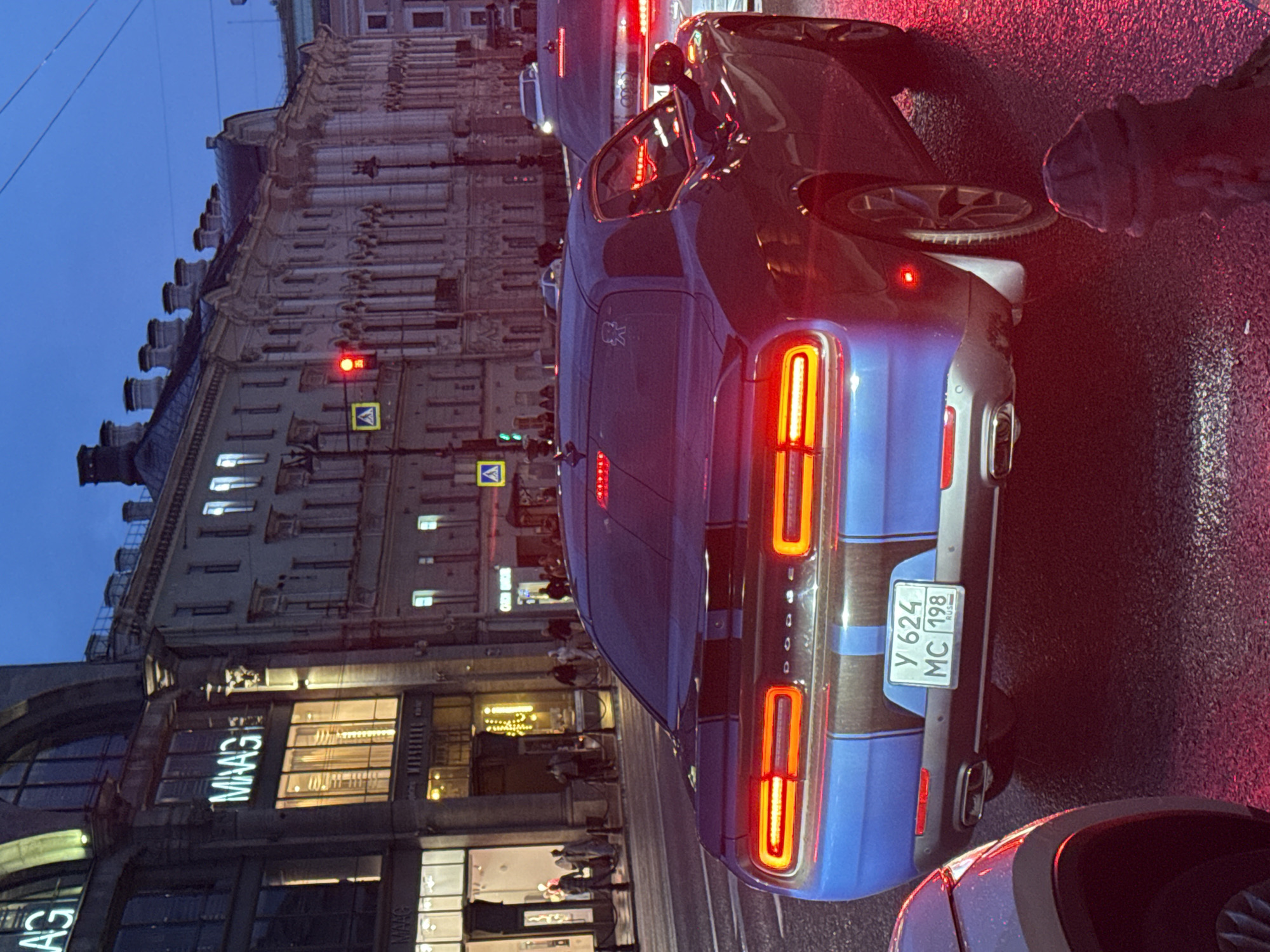 The image depicts a street scene in Saint Petersburg, Russia, likely during the evening or twilight hours, given the artificial lighting and the dim natural light. The focus is on the rear of a blue Dodge Challenger, which is stopped at a traffic light. The car has its brake lights illuminated, casting a red glow on the wet asphalt. The license plate is visible and reads "Y 624 MC 198" with "RUS" underneath, indicating Russian registration.
In the background, several multi-story buildings line the street, exhibiting architectural styles typical of Saint Petersburg, with ornate facades and numerous windows. One building on the left prominently displays the illuminated "MAAG" logo, suggesting it is a retail establishment. Pedestrians can be seen walking on the sidewalk in front of the shops. Traffic lights are visible, with one showing red, indicating the reason for the stopped vehicles. The overall environment suggests an urban setting with active street life.