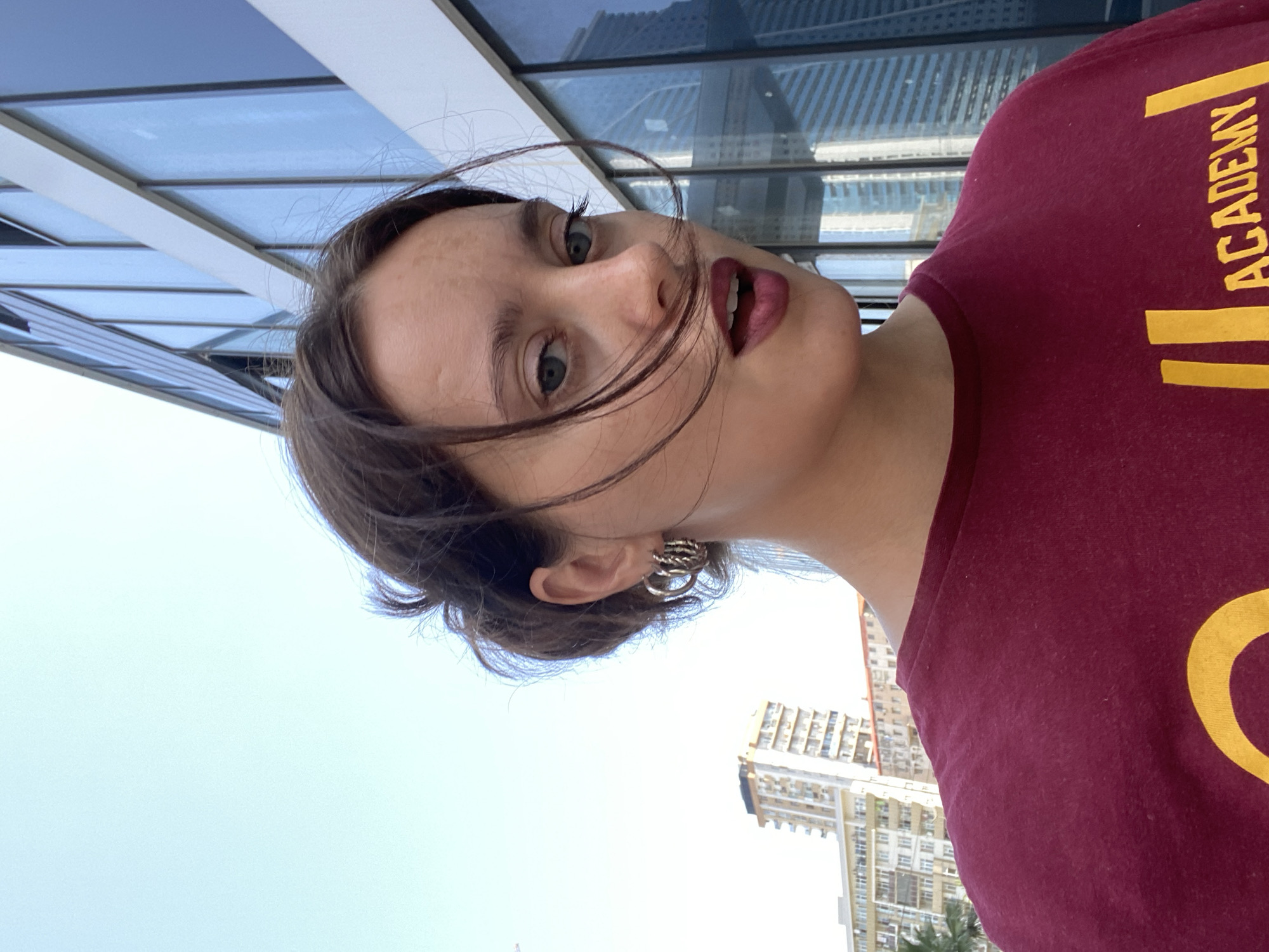 A young woman with dark hair pulled back in a bun and subtle freckles across her nose is shown in a close-up, outdoor selfie. She is wearing a maroon t-shirt with a yellow logo partially visible on the front. The t-shirt appears to have the word "ACADEMY" on it. She has large hoop earrings in her ears and is wearing dark lipstick. Her hair is windswept, with strands falling across her face.
In the background, a modern glass-fronted building dominates the right side of the frame. To the left of the building, several residential buildings are visible, appearing to be apartment blocks. One building, taller than the others, has a distinct facade with balconies. Palm trees are also visible in the distance. The sky is clear and bright blue. The scene suggests an urban environment in Batumi, Georgia, likely a sunny day.