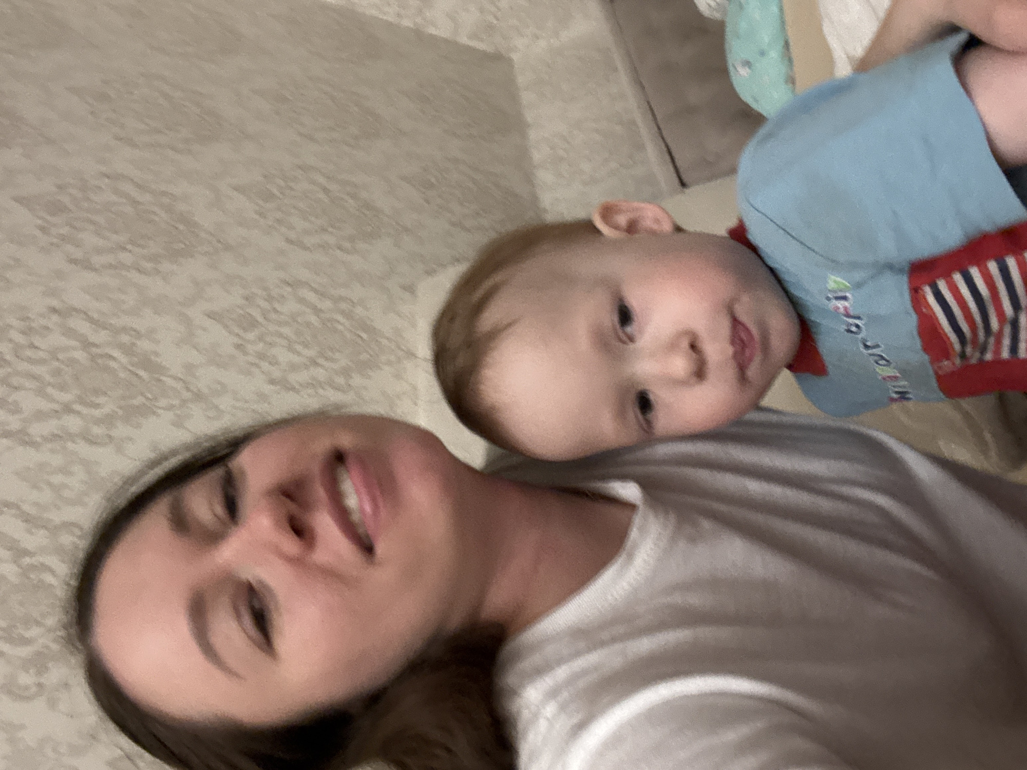 The image shows a woman and a young child. The woman has long brown hair and is wearing a white t-shirt. The child is wearing a blue and red shirt. They appear to be lying down, possibly taking a selfie. The background is a textured wall, which might indicate an indoor setting. The location is Ферма, Russia.