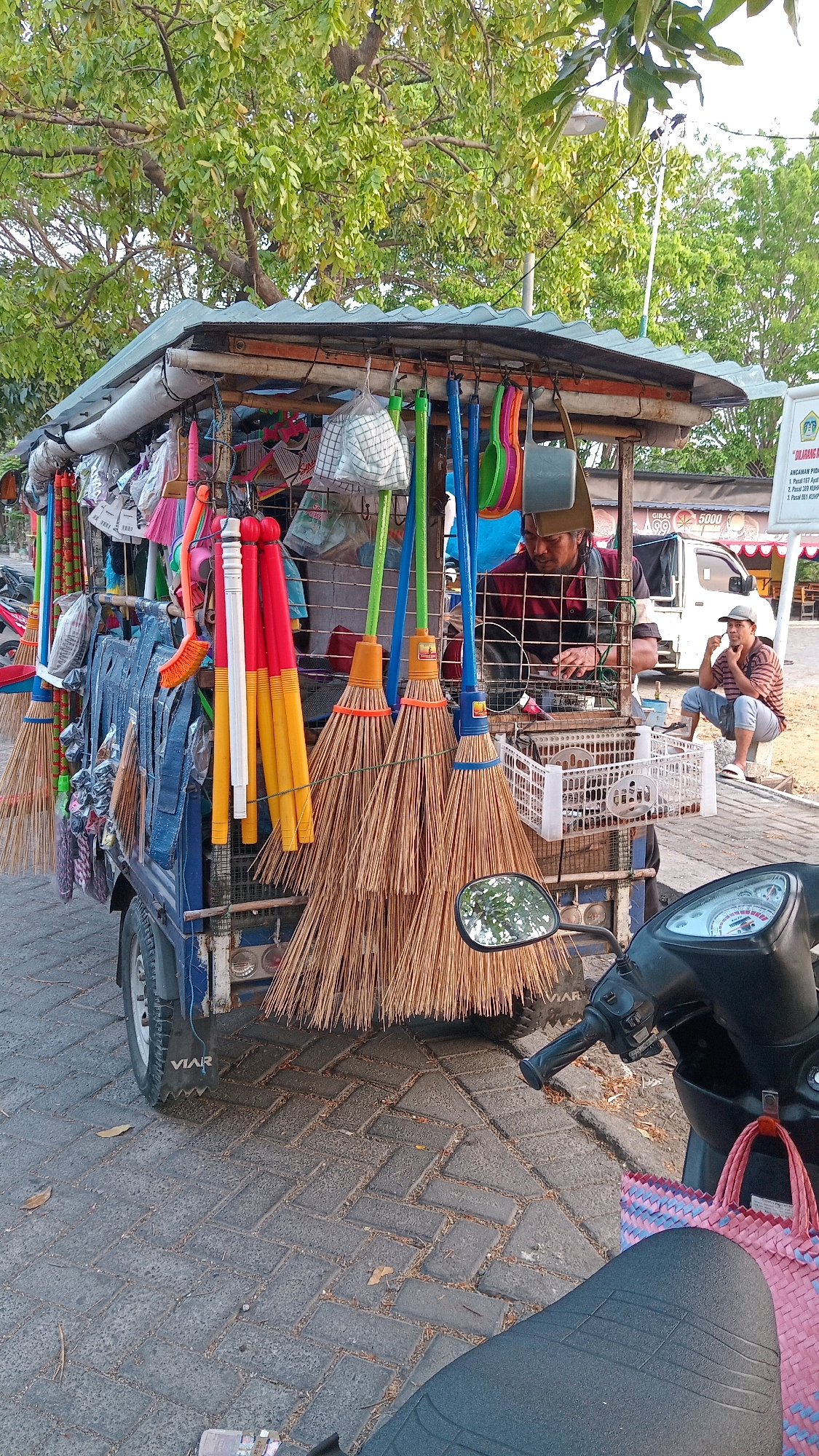 This image depicts a street vendor's cart filled with brooms and other cleaning supplies, located in Pongangan, Indonesia. The cart, a blue motorized vehicle with a makeshift canopy, is laden with a variety of brooms made of natural fibers. Hanging from the canopy are also several colorful plastic buckets and dustpans in shades of green, blue, and pink. Behind the cart, two men are visible. One man, wearing a red and black checkered shirt, appears to be attending to the cart's contents. The second man, seated and wearing a striped shirt, is observing. The scene is set on a paved street, with trees and vegetation visible in the background, suggesting an outdoor setting. A motorcycle is parked in the foreground, with a pink woven bag resting on its seat.
