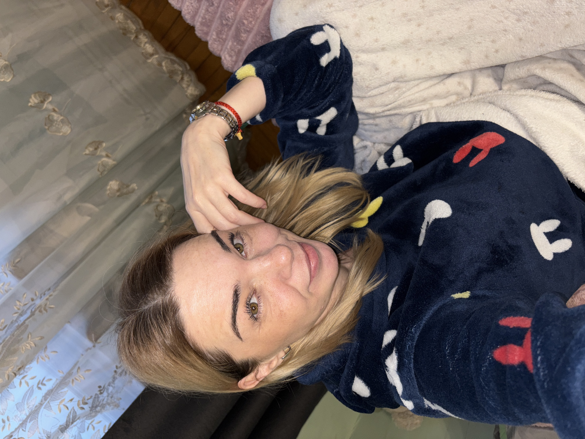 A woman with blonde hair is lying on a bed, wearing a navy blue pajama top with a pattern of red and white shapes. She is looking directly at the camera with a slight smile. Her left hand is raised, resting on her head near her hairline. A watch and a red bracelet are visible on her left wrist. The bedding consists of a white blanket with a subtle star pattern and a pink, textured throw at the edge of the frame. To the left of the woman, a sheer curtain with an embroidered floral design partially covers a window, casting soft light on the scene. The location is unknown.