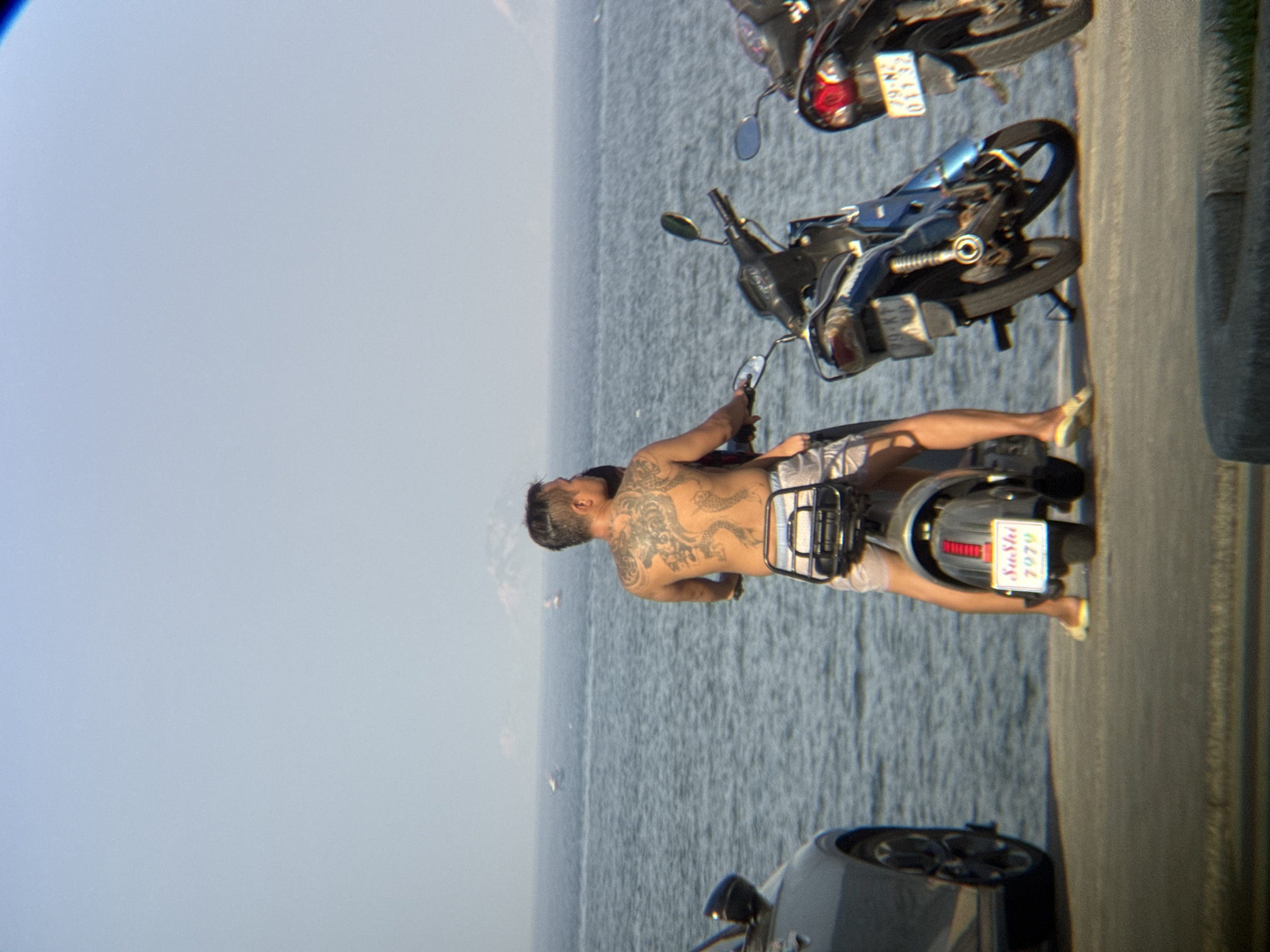 The image depicts a sunny day scene near the coast of Nha Trang, Vietnam. In the foreground, a man with extensive back tattoos is seated on a scooter, facing away from the camera. He is wearing light-colored shorts. To his left, the rear section of a silver car is visible, parked near the water's edge. Behind the man and his scooter, a body of water, presumably the sea, stretches out under a clear sky. In the distance, several small boats are scattered across the water. To the right of the main scooter, another scooter is parked, and beyond that, a portion of a third scooter is visible. The ground is paved, suggesting a promenade or parking area adjacent to the beach. The overall atmosphere is relaxed and casual, indicative of a typical seaside location.