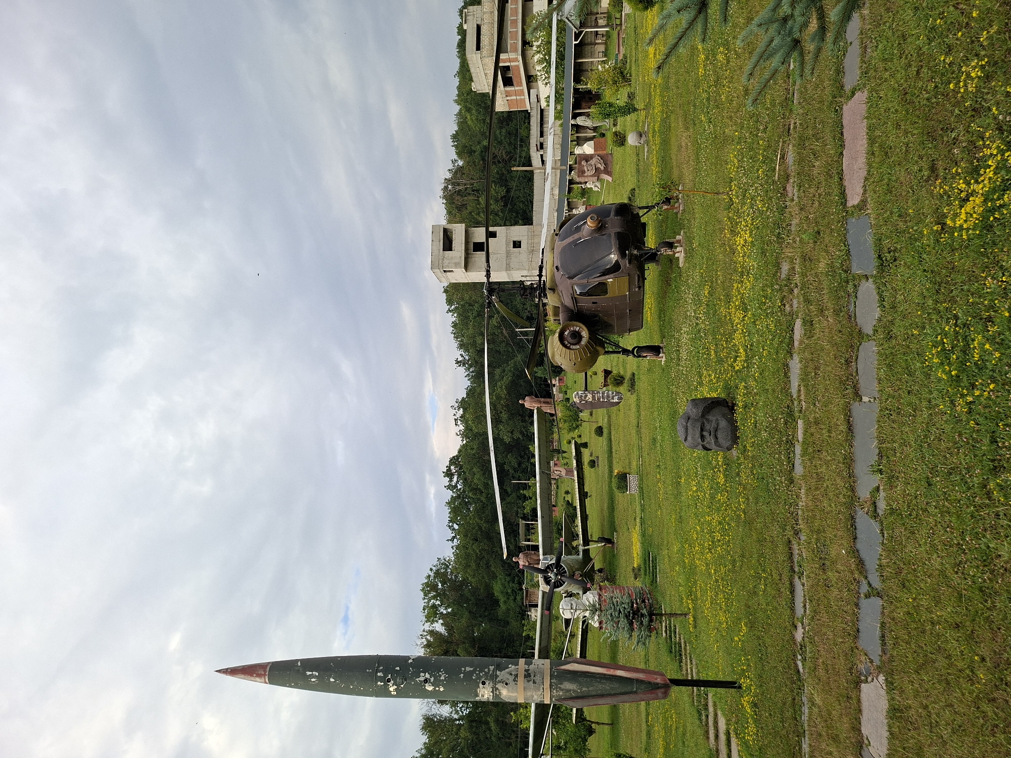 This is a medium-shot, eye-level outdoor photograph taken in the city of Malcoci, Moldova. The scene is an open field with scattered objects, primarily military artifacts and sculptures, under a cloudy sky. In the foreground, a dark stone bust of a man with a beard is positioned on the grass. To the left, a large, dark green missile with a red tip stands upright. Behind the missile, a vintage airplane with its propeller visible is parked on the grass. Further back and to the right, a dark brown and green helicopter is also on display. Several statues of people are scattered throughout the field, along with some small evergreen trees and bushes. In the background, unfinished concrete buildings rise above a line of trees. The field is covered in green grass with numerous small yellow flowers. A pathway made of stone slabs runs through the foreground, leading towards the right side of the frame.