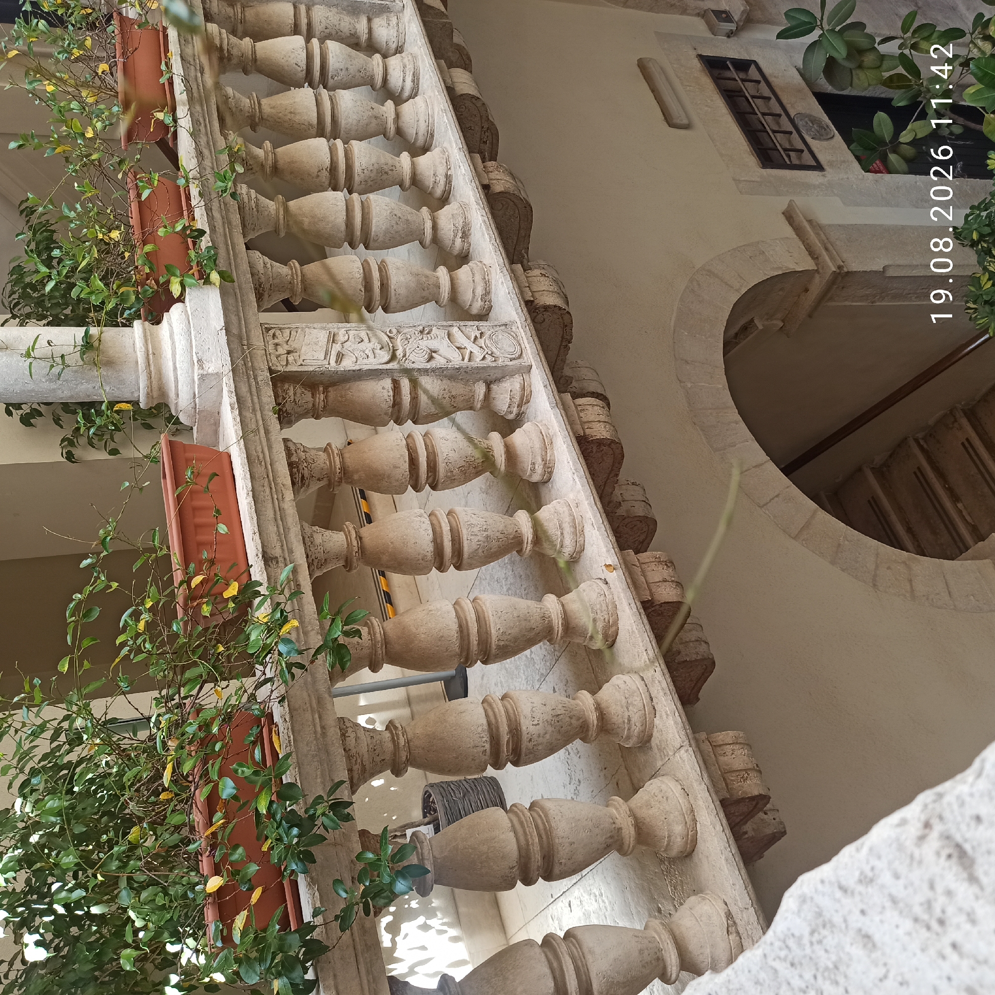The image depicts a balcony with a stone balustrade and planters filled with green plants. The balustrade features decorative carvings and turned stone posts. Below the balcony, an arched entryway leads to a staircase. The architectural style suggests an older building, possibly in a historic district. The timestamp "19.08.2026 11:42" is overlaid on the bottom right of the image. The location is identified as Ruvo di Puglia, Italy. There are no people visible in the image.
