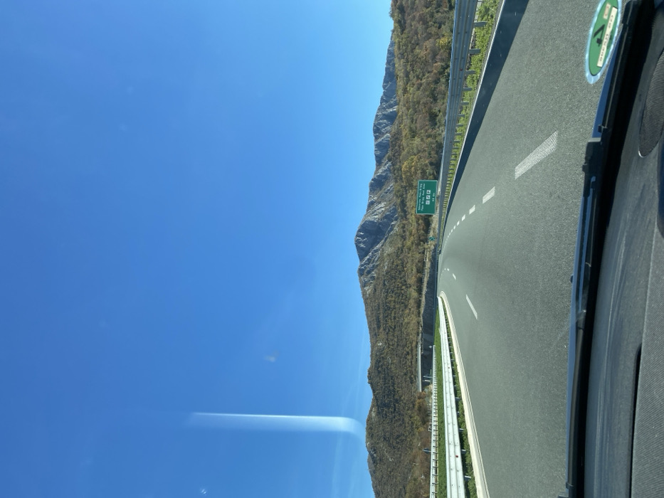 The image displays a multi-lane asphalt highway on a clear, sunny day, viewed from the interior of a vehicle traveling towards Pelev Brijeg, Montenegro. The sky, a deep clear blue, occupies the upper half of the frame, with a prominent vertical light streak or reflection visible on the left side of the windshield.
In the mid-ground, a range of mountains stretches across the horizon. The mountains on the left are covered with vegetation exhibiting autumnal or dry tones, while the mountains on the right feature more exposed, rugged grey rock faces.
The highway comprises at least two lanes in the direction of travel, marked by white dashed lines. On the left, a white metal guardrail follows the curve of the road, and a concrete overpass or bridge structure is visible in the distance. On the right, another white metal guardrail borders the road, beyond which is a vegetated slope leading to the mountains.
A green overhead road sign is positioned on the right side of the highway. It indicates "Montenegro Pelev Brijeg" in Latin script, along with Cyrillic text, symbols for a gas station and restaurant, and the distance "2.4 KM".
The foreground shows the black dashboard and the lower edge of the vehicle's windshield. A windshield wiper blade is partially visible, and a circular green and white sticker is affixed to the lower right portion of the windshield. No individuals are visible in the scene.