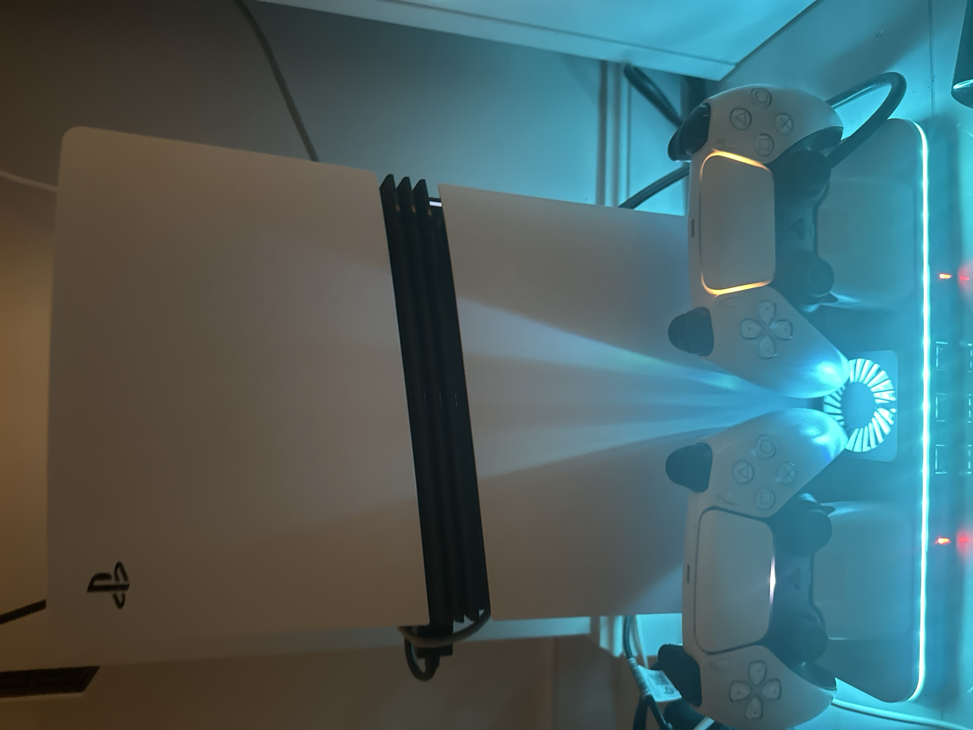 The image displays a modern gaming setup featuring a white PlayStation 5 console and two white DualSense controllers, illuminated by a cool blue-teal light. The PlayStation 5 console is positioned vertically, showcasing its distinctive white side panels and the black central section. A neatly bundled set of black cables runs along the left side of the console. The iconic PlayStation logo is visible on the lower left portion of the console's front panel.
To the right of the console, two white DualSense controllers are docked vertically on a charging station. This station incorporates a circular cooling fan, visible behind the lower controller, and a prominent vertical RGB light strip along its right edge, which is the primary source of the blue-teal illumination in the scene. The light bars on both controllers are glowing in an orange-yellow hue, indicating they are actively charging.
The overall environment is dimly lit, suggesting an indoor setting during the evening or night, with the artificial blue light creating a distinct ambiance. The setup appears to be placed against a light-colored wall or shelf. The location provided for this media is Khimki, Russia.