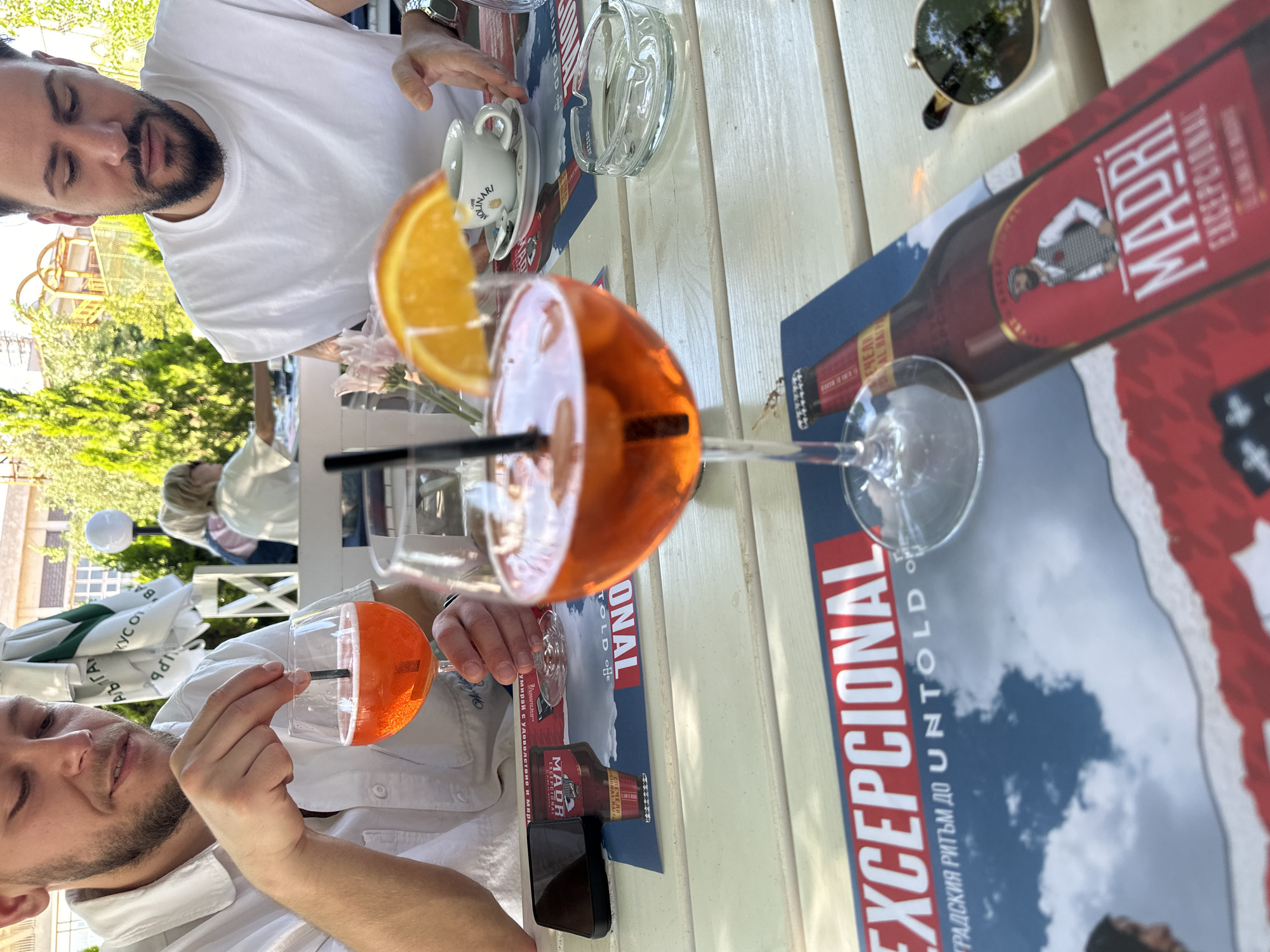 Two men sit at a white table outdoors. Both are wearing white shirts. In front of them are Aperol Spritz cocktails in tall glasses garnished with orange slices and black straws. An ashtray, a teacup, and sunglasses sit on the table. There are Madri Exceptional beer advertisements on the table. There are trees and buildings visible in the background. Based on the Cyrillic alphabet on one of the advertisements, the location may be in Bulgaria, possibly Plovdiv due to the presence of a local sign.