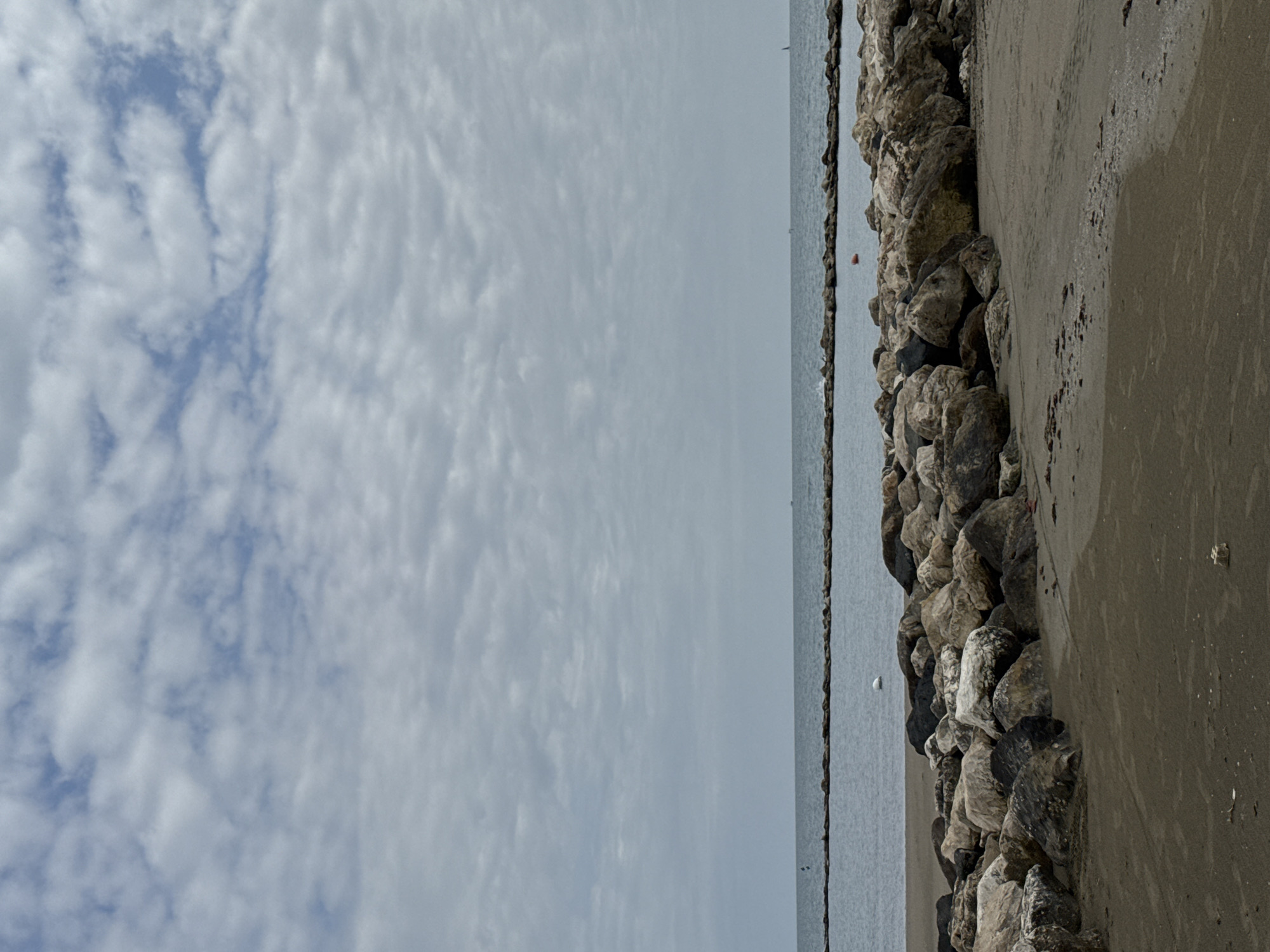 The image displays a coastal scene under a cloudy sky. The horizon line is visible, separating the expanse of the sky from the calm sea.
In the upper portion of the frame (when correctly oriented), a sky filled with various formations of white and grey clouds dominates, with patches of light blue sky visible through thinner areas. The clouds appear diffuse, suggesting an overcast or partly cloudy day.
Below the sky, the sea stretches horizontally across the middle of the image. The water surface appears relatively still and greyish-blue, reflecting the muted light from the sky. A few small, indistinct objects, possibly distant boats or buoys, can be seen on the water's surface, and one prominent red buoy is visible closer to the shore.
The lower portion of the image features the shoreline, composed of a line of large, rugged rocks forming what appears to be a breakwater or natural barrier. These rocks vary in size and color, from light grey to darker tones, and are covered in some growth or stains. Immediately adjacent to the rocks on the right side is a strip of sandy or dirty ground, textured and uneven, with some darker patches and small debris. No people are visible, and there is no specific activity taking place. The overall impression is one of a quiet, somewhat serene coastal environment during daylight hours. No visible text is present. Given the context, this scene is located in Nettuno, Italy.