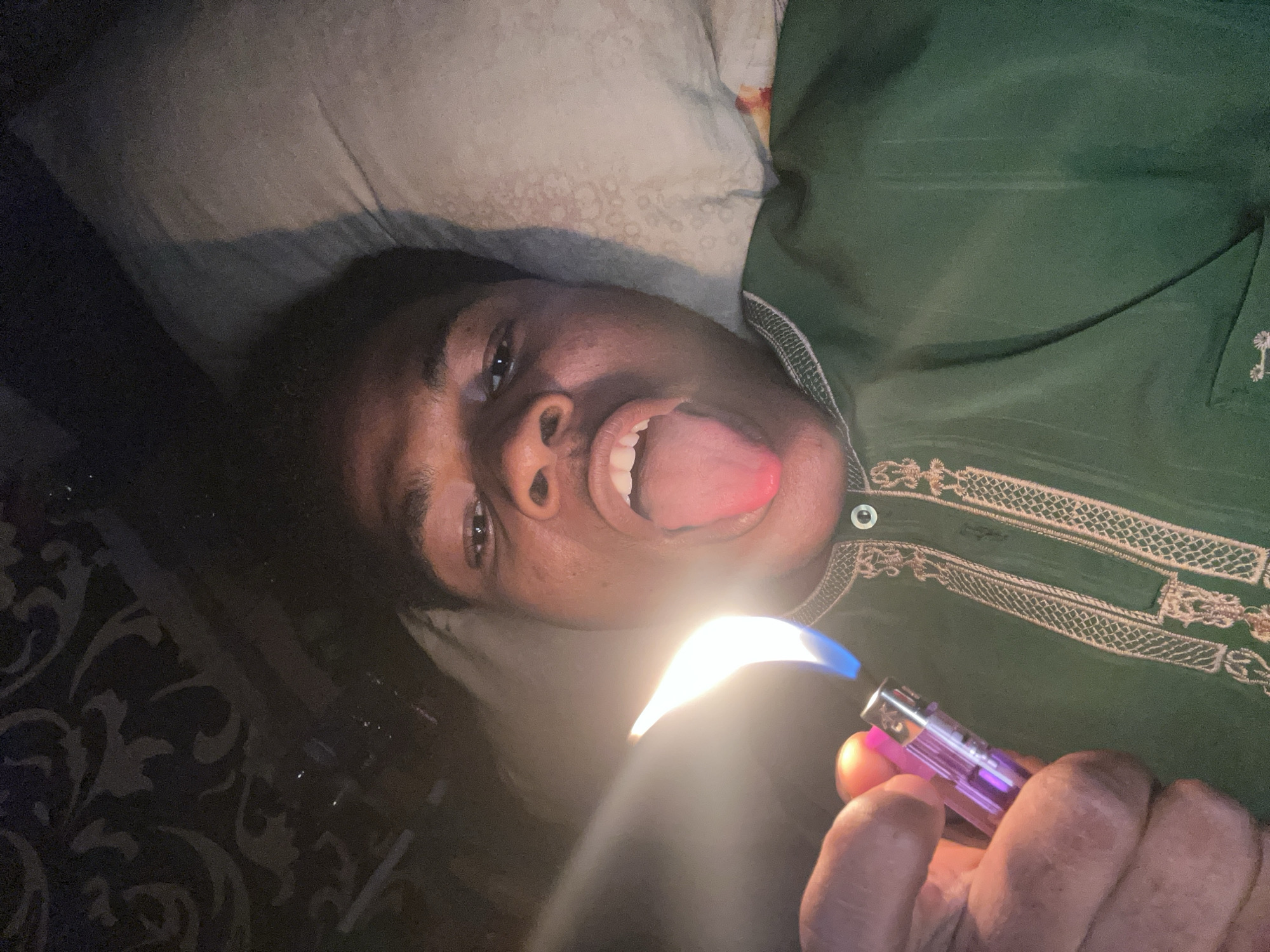 The media file shows a close-up, overhead shot of a young person lying down, holding a pink lighter with the flame ignited. The person is wearing a green garment with intricate embroidery around the neckline and down the front. Their head is tilted back, resting on a pillow, and their mouth is open with their tongue sticking out, facing the viewer. The flame from the lighter is bright, casting a strong light on the person's face and obscuring part of their chin and nose. The background consists of a patterned dark fabric and a pillow. The overall lighting suggests it is likely nighttime or indoors. The presence of a lighter and the person's expression could indicate a moment of playfulness or experimentation. Given the location context of Ihiagwa, Nigeria, the scene is set within a domestic or personal space in that region. There is no visible text in the image.