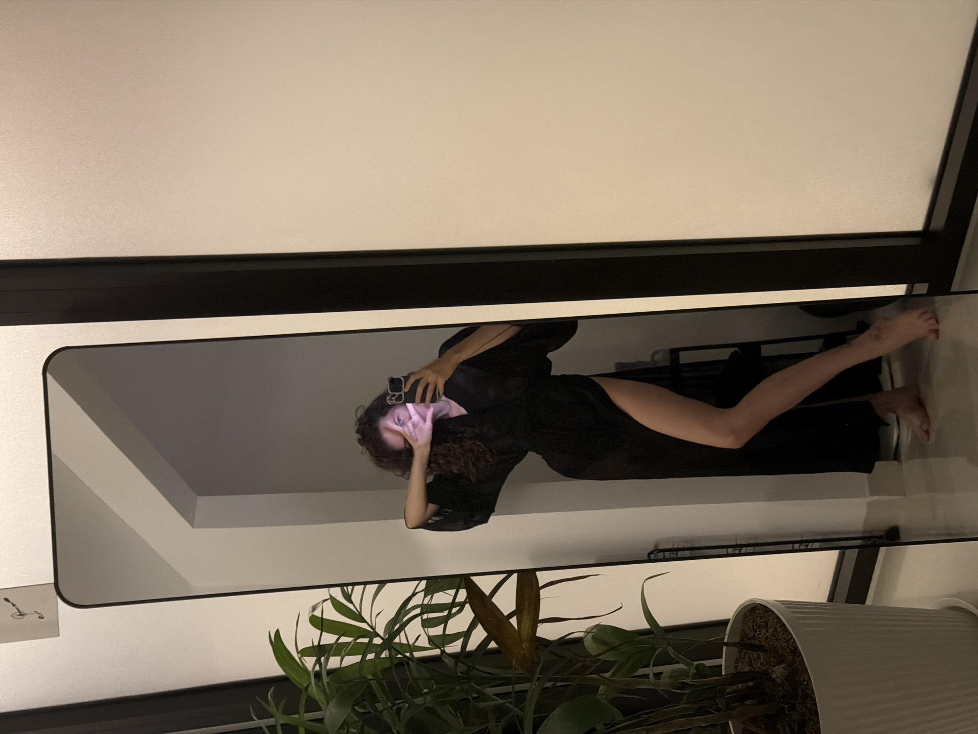 The image, taken in Đà Nẵng, Vietnam, depicts a person taking a mirror selfie indoors during what appears to be the evening or night. The main subject is a woman, reflected in a large, floor-length, rectangular mirror with a thin dark frame. She is wearing a dark, possibly sheer, long-sleeved garment that resembles a robe, with her left leg visible.
The woman is holding a black smartphone with her right hand to capture the selfie, while her left hand is positioned near her face, partially obscuring it, with fingers forming a "V" or peace sign gesture. Her curly dark hair is visible around her head and shoulders.
The setting is an indoor space with light-colored walls, possibly white. A dark horizontal structure, possibly a window frame or a shelf, runs across the upper part of the background. In the foreground, at the bottom of the mirror's reflection, is a potted plant with green foliage, indicating an indoor plant. The artificial lighting creates a warm, subdued ambiance. No discernible text is visible in the image.