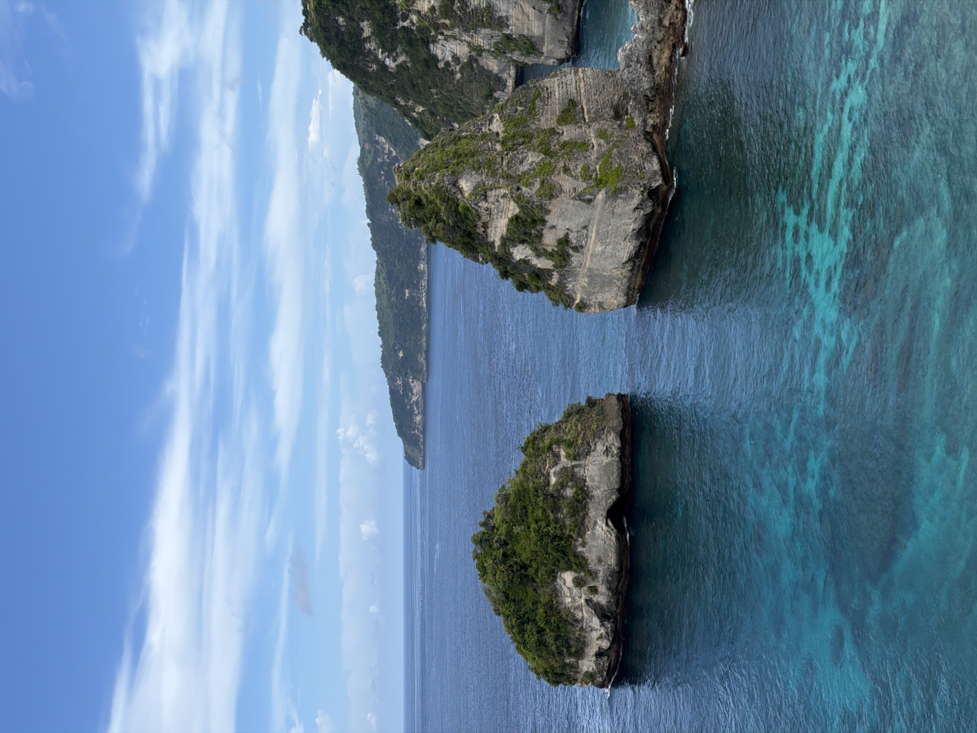 The image captures a breathtaking natural seascape in Nusa Penida, Indonesia, featuring dramatic rocky formations emerging from clear, vibrant waters under a bright sky. The perspective appears to be from a higher vantage point, looking out over the ocean.
In the foreground and midground, two prominent rocky islets stand majestically in the sea. These islets are rugged, grey rock formations, heavily adorned with lush green vegetation on their tops and upper slopes, suggesting a verdant, tropical environment. The water surrounding them is a mesmerizing blend of deep blue, transitioning to vivid turquoise closer to the rocks, with discernible lighter patterns beneath the surface indicating shallow areas or coral formations.
In the distance, a larger, elongated landmass, likely a part of the island's coastline, stretches across the horizon. This landmass also appears to be covered in dense green foliage, adding to the remote and natural feel of the scene. The sky above is a clear, bright blue, accented by delicate, wispy white clouds that extend across the frame.
The image portrays a serene and pristine natural environment during daytime, with excellent visibility. There are no signs of human activity, structures, or visible text, highlighting the untouched beauty of this Indonesian coastal landscape.
