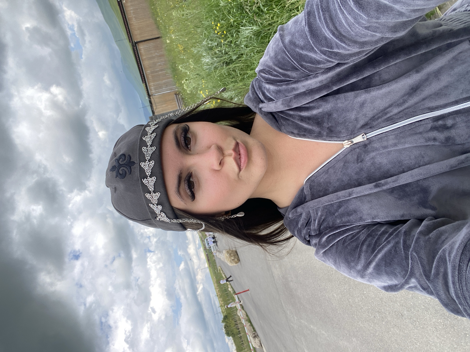A young woman with dark hair, wearing a grey velour tracksuit and a decorative grey cap with a chain and earring, takes a selfie outdoors. She is facing the camera with a slightly pursed-lip expression. Behind her, a paved path stretches into the distance, bordered by green grass, some trees, and a wooden fence. Several people are visible in the background walking on the path. The sky is cloudy and grey, with mountains visible in the distance. The location appears to be in Tsalkita, Russia.