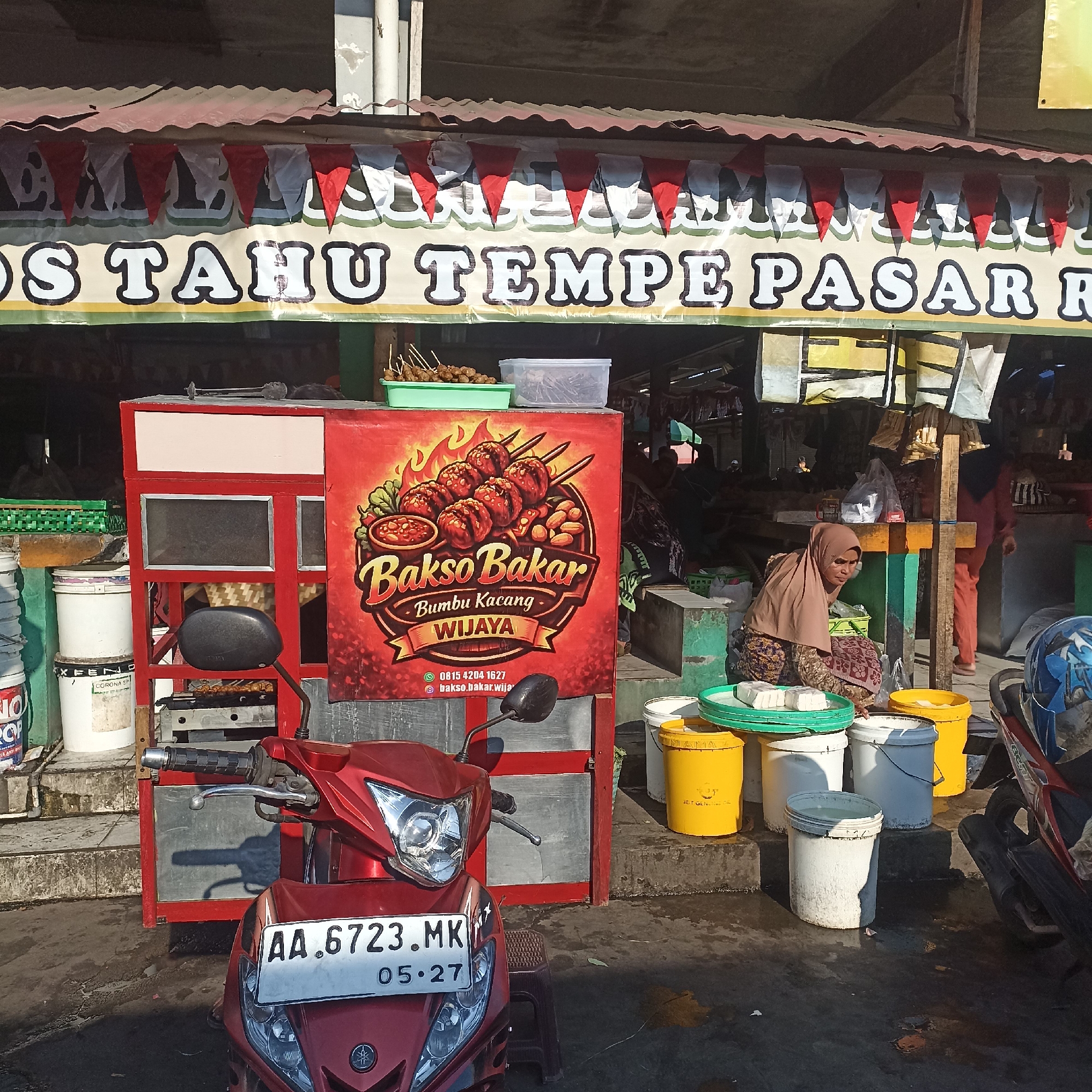 The image depicts an outdoor market scene in Magelang, Indonesia, likely on a sunny day given the shadows. In the foreground, a red motorcycle with license plate "AA. 6723. MK" is parked, facing left. To the left of the motorcycle is a food stall with a red and white structure, displaying a banner that reads "TAHU TEMPE PASAR RA" with red and white triangular flags above it. A grill with food is visible underneath the stall. To the right of the stall, a woman wearing a brown hijab and a patterned skirt is crouched down, tending to several large buckets. Some of the buckets contain white blocks, possibly tofu, on top. There are also various other buckets, some yellow and white, arranged around the stall and the woman. In the background, more market stalls and people can be seen, suggesting a bustling commercial area. The ground is wet and appears to be paved.