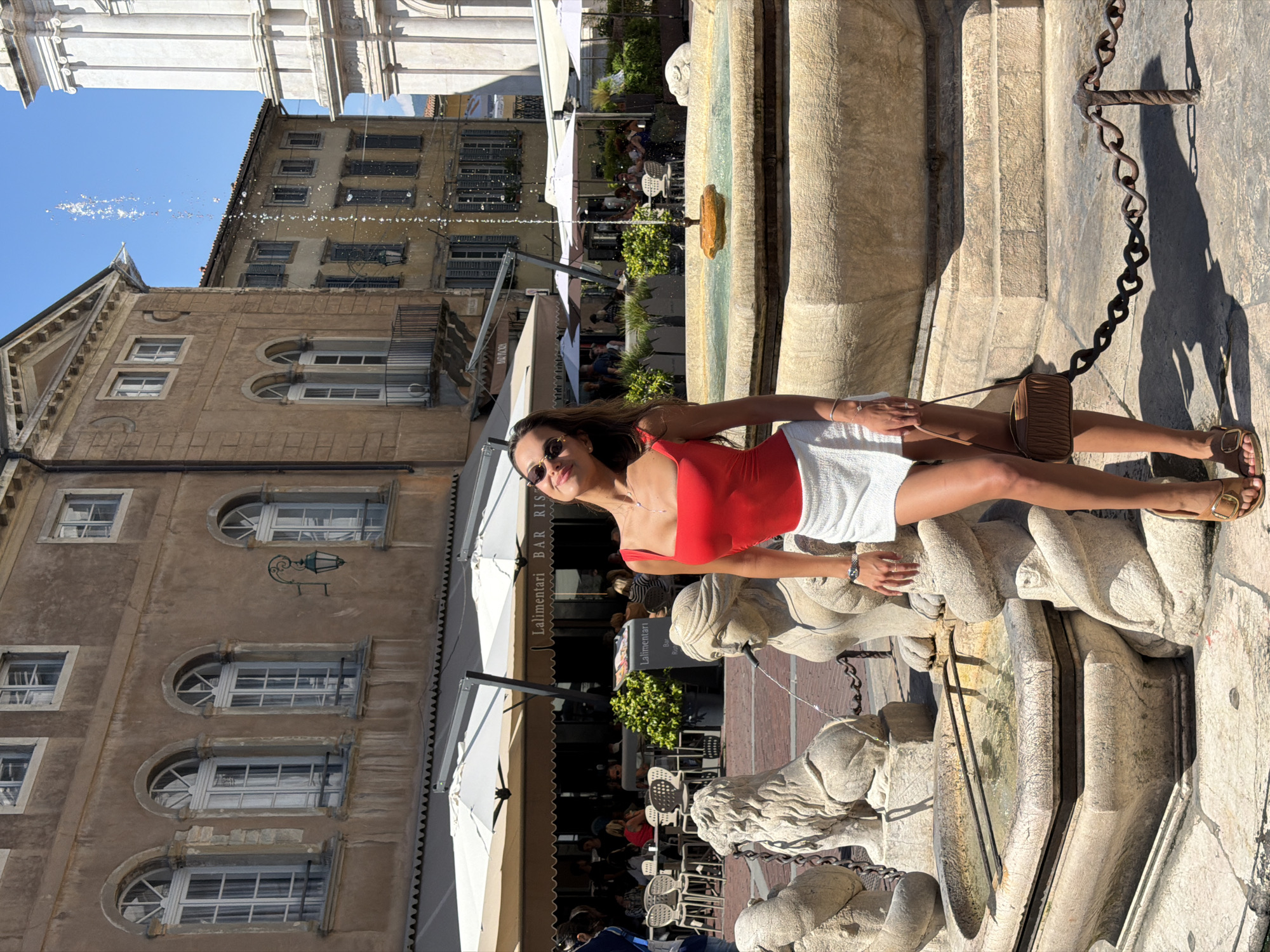 A woman poses in front of a fountain in Mozzo, Italy. The woman is wearing a red tank top and a white skirt. She is holding a brown purse and wearing brown sandals. The fountain is made of stone and has statues of lions. In the background, there are buildings with shops and outdoor seating. The sky is blue and there are some clouds. The woman is smiling and looking at the camera. The scene is brightly lit by the sun.