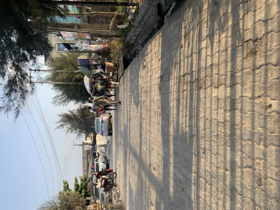 The image depicts a street scene in Ikota, Nigeria, under clear daylight, casting long shadows indicative of morning or late afternoon. The foreground features a wide, paved road constructed of interlocking concrete blocks, showing signs of dirt and wear.
On the left side of the road, a person in a light-colored shirt is seated on a brown motorcycle. Nearby, another individual in a dark dress stands on the paved surface. Behind them, multiple vehicles are visible, including a silver SUV and a white sedan, parked amidst piles of wooden pallets and construction debris. Utility poles with overhead power lines are present on this side.
In the center of the road, a silver sedan is either parked or moving slowly away from the camera. A group of three individuals walks away from the camera in the middle-right of the paved area; one wears a striped shirt, another a patterned garment.
The right side of the street features a series of makeshift market stalls operating under two distinct, colorful umbrellas (one white and green, one blue and white). Various goods, including baskets and produce, are displayed. A narrow, concrete-lined drainage ditch runs parallel to the road, containing dark liquid and refuse. A rudimentary wooden plank bridge crosses this ditch to access one of the stalls. Behind the stalls, a tall fence constructed of bamboo or similar reeds is visible, adorned with several banners or posters. A multi-story building with light-colored walls and blue-framed windows stands further back. Tall, slender trees with sparse foliage line both sides of the street, and additional utility poles with power lines are present on the right.