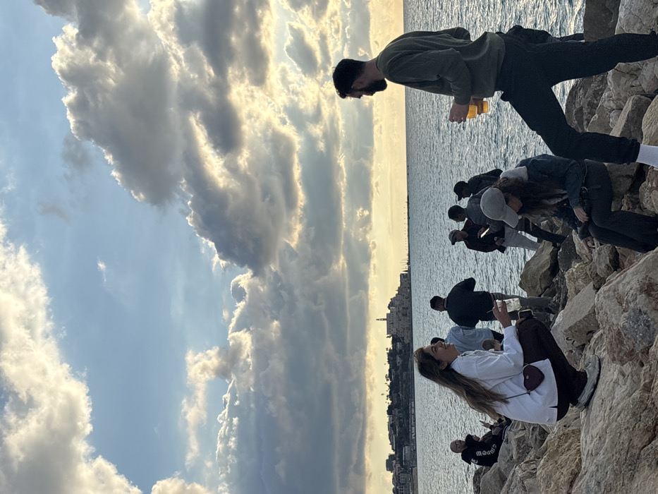 The image depicts a group of individuals gathered on a rocky shoreline of the Mediterranean Sea in Tel Aviv, Israel, during late afternoon or sunset. The historic cityscape of Jaffa (Yafo), characterized by its traditional buildings and a prominent clock tower, is visible across the water in the distant background. The sky features large cumulus clouds, backlit by the low sun, creating luminous edges and scattered light. The sea surface is rippled and dark blue-grey.
In the immediate foreground, a female with long brown hair, wearing a white long-sleeved shirt and dark pants, is crouched on the rocks, holding a clear plastic cup. A small, reddish-brown heart-shaped purse is slung over her shoulder. To her right, a second individual, wearing a grey baseball cap and a dark denim jacket, is seated on the rocks, looking downwards. A bearded male, wearing an olive-green long-sleeved top and dark pants, stands to the far right, holding two clear glasses containing an amber liquid. Approximately five to seven additional individuals, primarily male, dressed in casual clothing, are scattered in the mid-ground on the rocks. They are standing or seated, engaged in social interaction, with some observed holding cups. The scene portrays a casual outdoor social gathering by the sea.